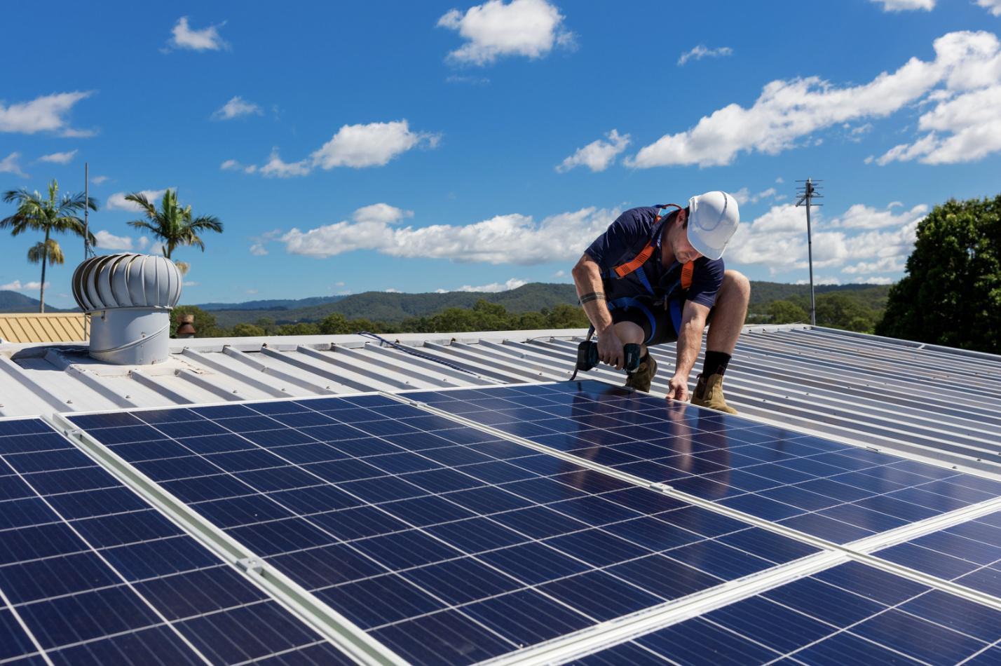 A worker installing solar panels on a rooftop under a blue sky with clouds.