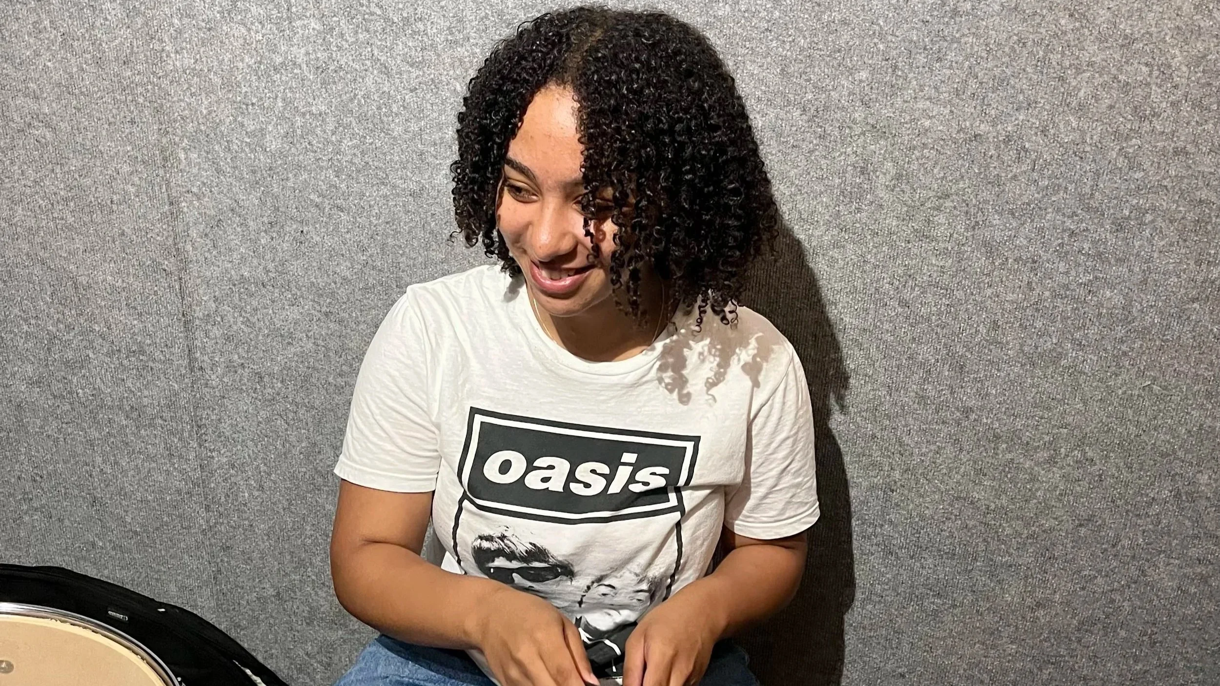 A young woman with curly black hair smiling while sitting against a gray wall, wearing a white Oasis band t-shirt and blue jeans.