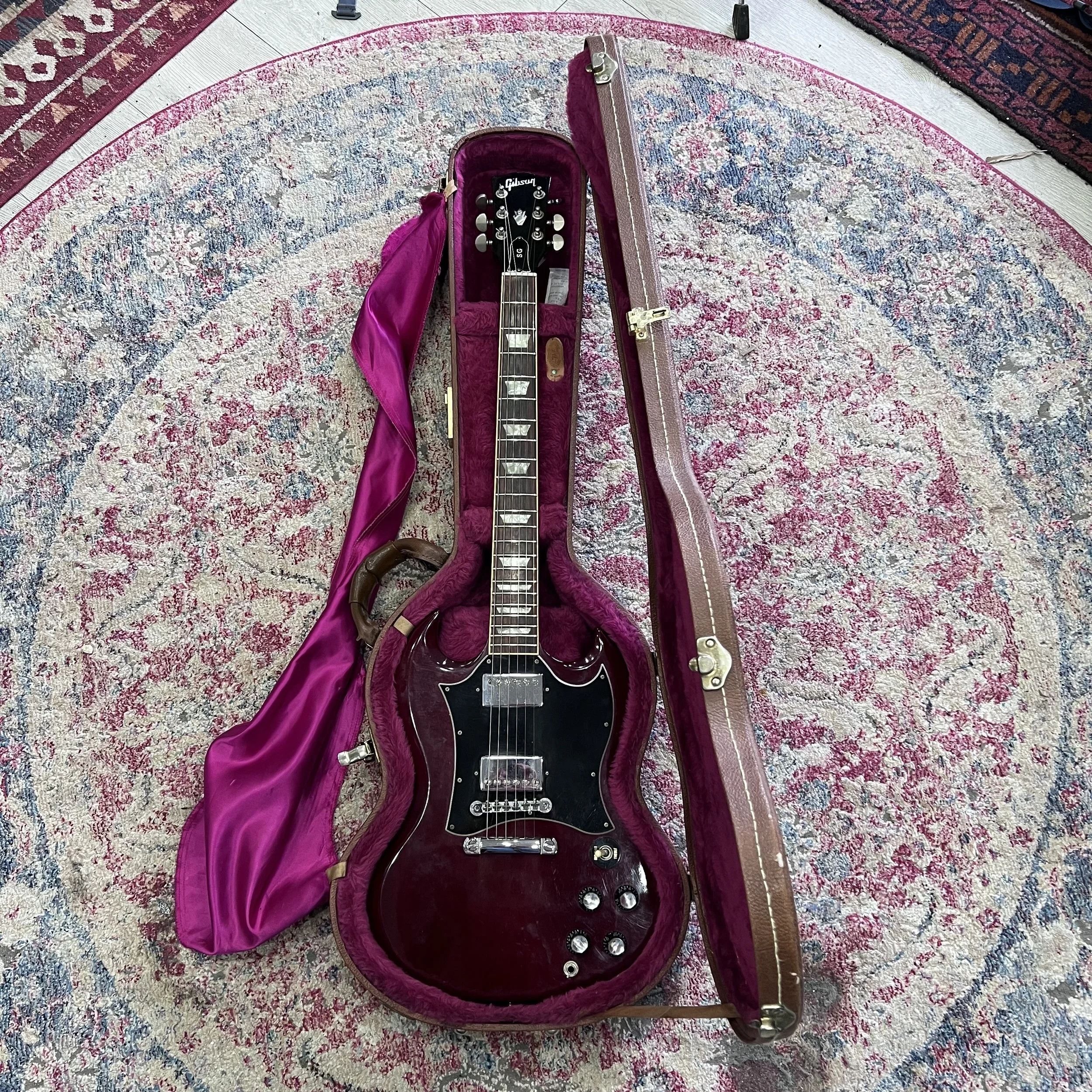 Gibson SG Standard electric guitar in a cherry finish, featuring a black pickguard, trapezoid inlays, and chrome humbuckers, resting in a vintage-style brown hard case.jpeg