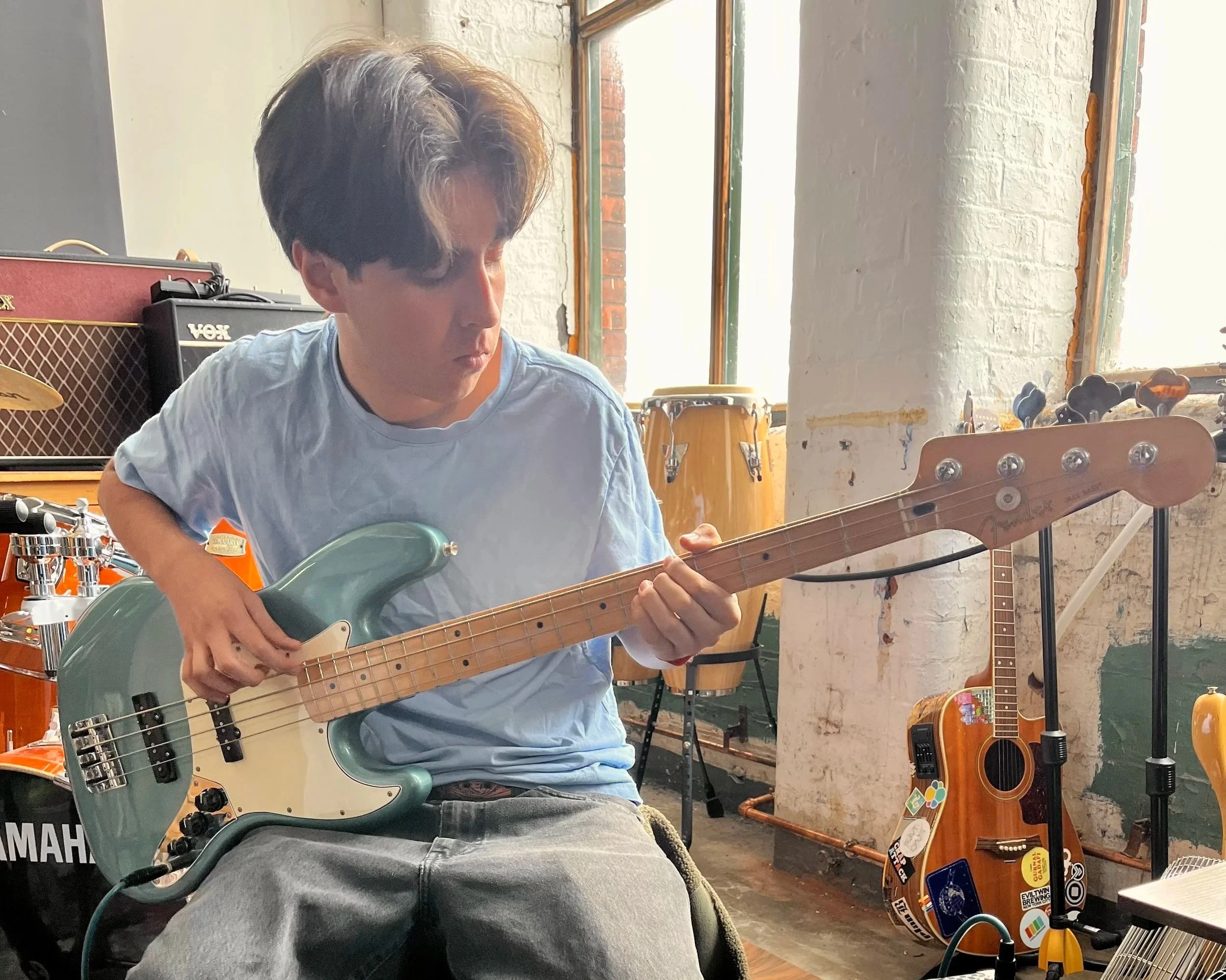 A young man with dark hair playing an electric bass guitar in a music studio. There are musical instruments and equipment like a guitar, drums, and amplifiers around him.