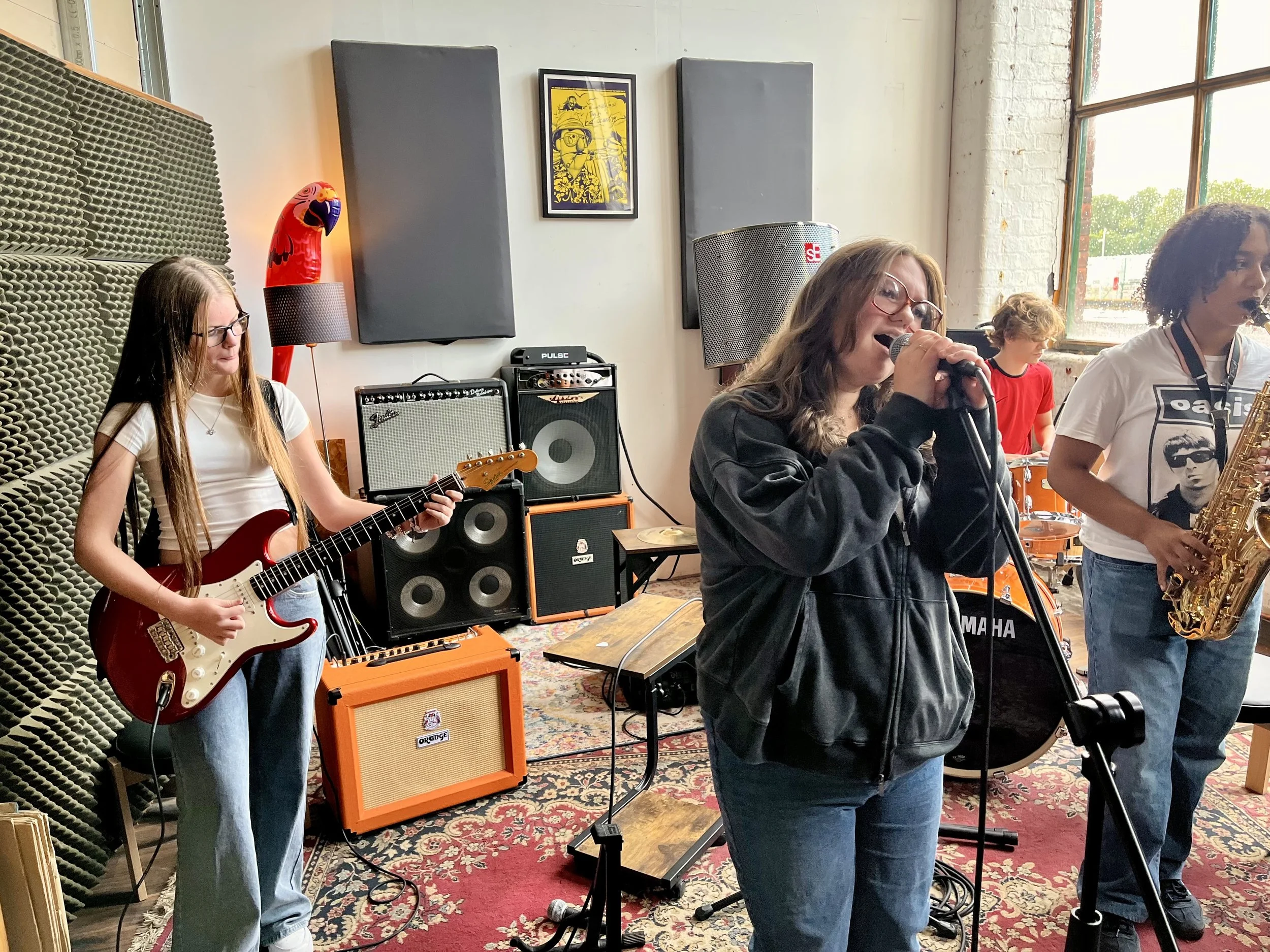 A girl with glasses playing an electric guitar on a stage with musical amplifiers, a woman singing into a microphone, a boy playing the drums, and a woman playing the saxophone, in a room with large windows and artwork on the wall.