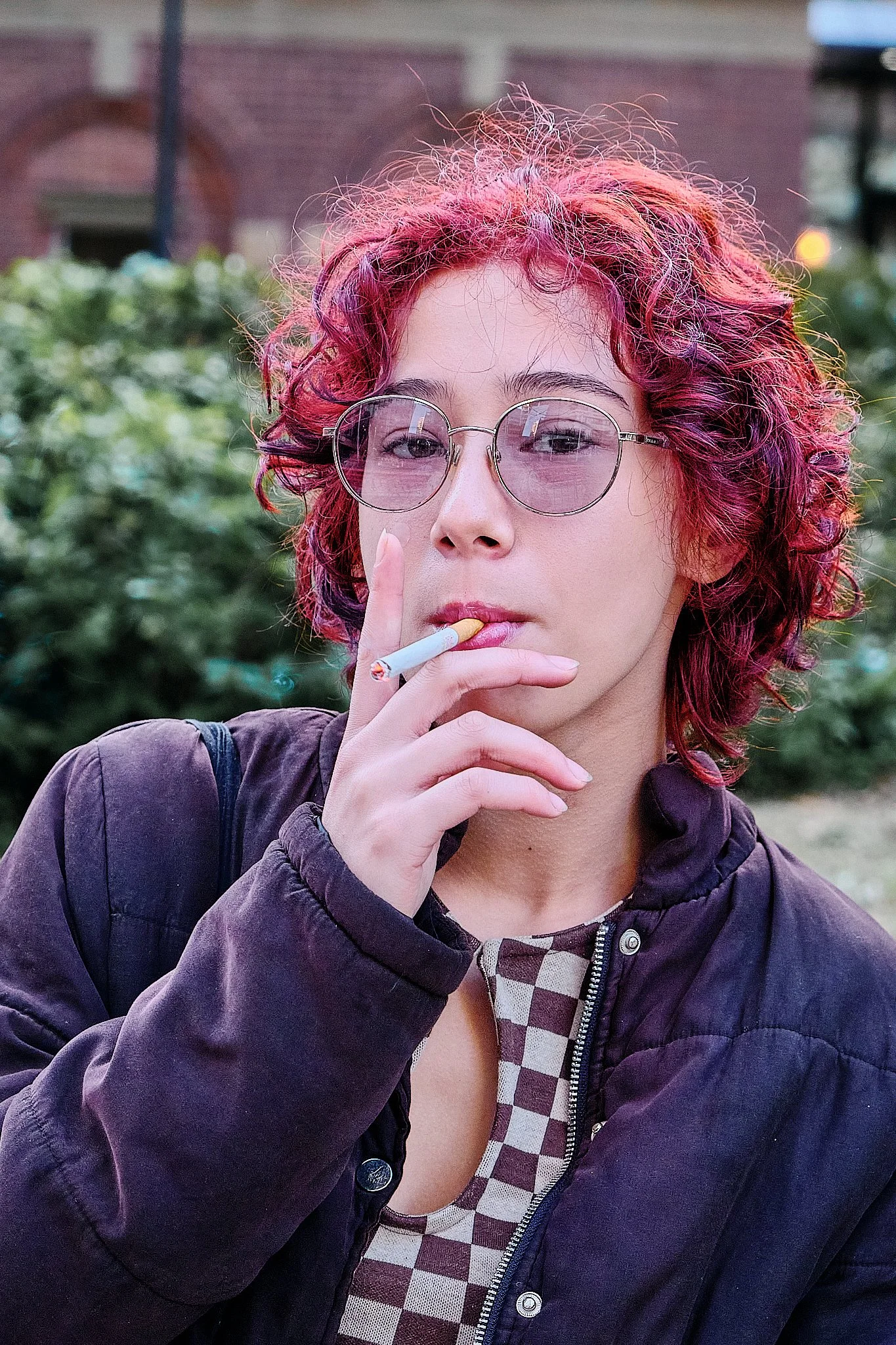 A woman with curly, red and purple hair wearing round sunglasses and a dark jacket, holding a lit cigarette near her lips, outdoors with greenery and a brick building in the background.