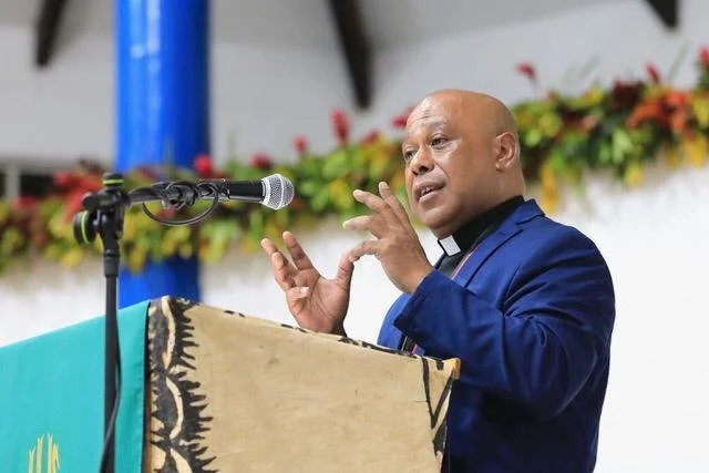 A man in a blue suit and clerical collar speaking at a podium with a microphone, with colorful flowers hanging on the wall behind him.