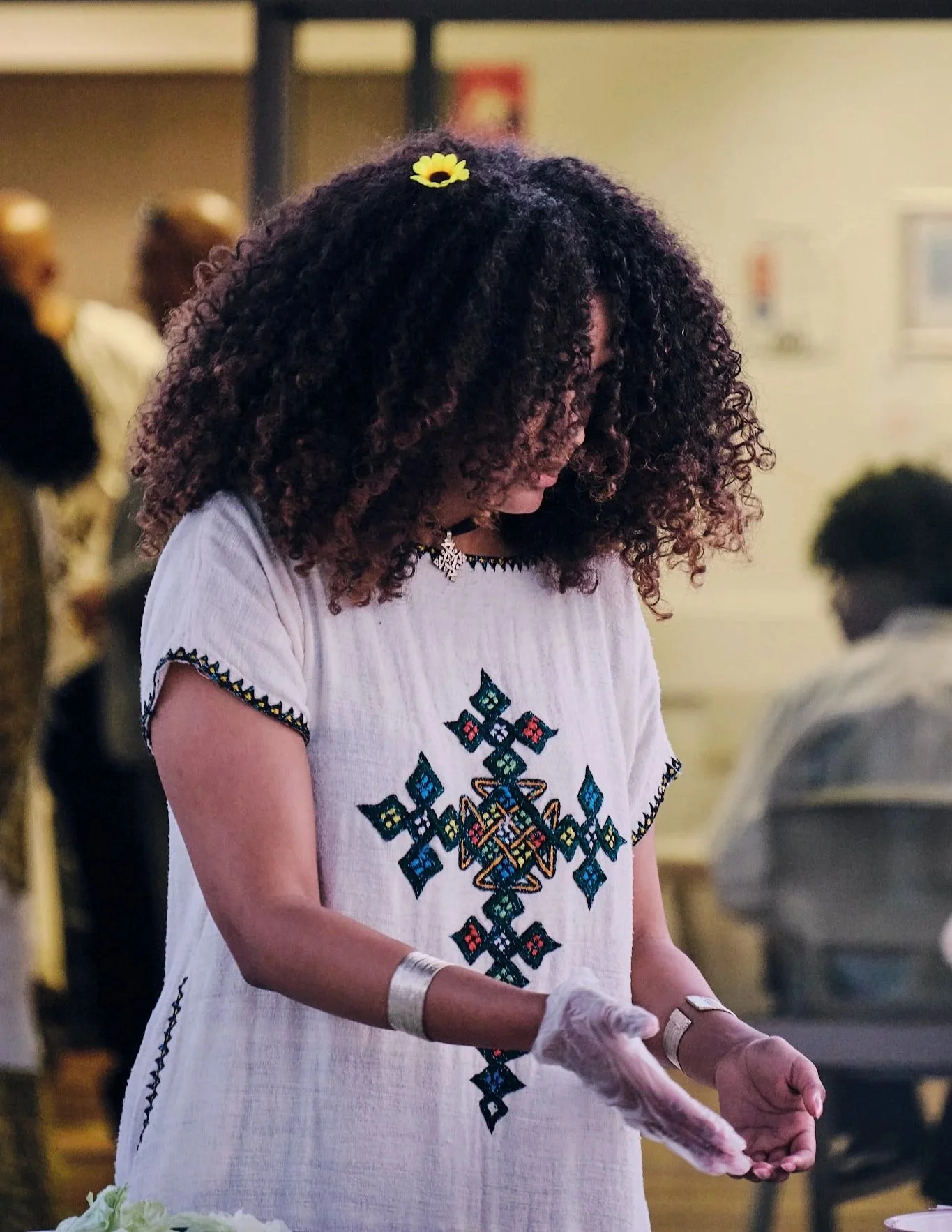 An Ethiopian Australian woman with curly hair wearing a traditional Ethaiopian shemma dress, with a sunflower in her hair, and silver jewelry, standing in a busy indoor setting.