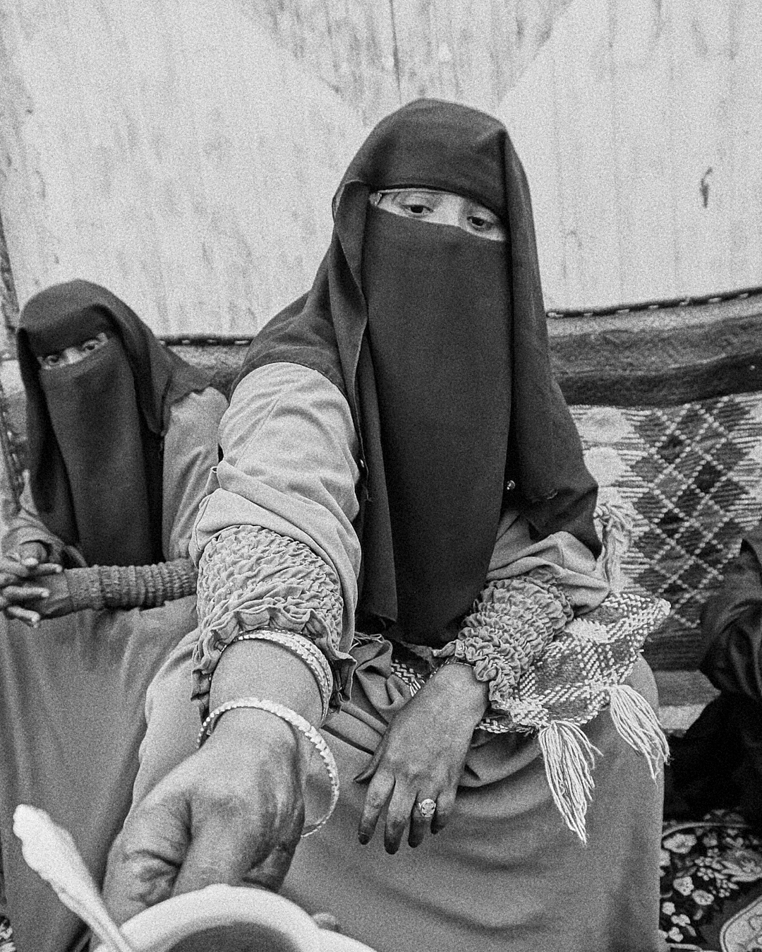 Two Afar women wearing niqabs and traditional clothing, sitting indoors with rugs and blankets around them.
