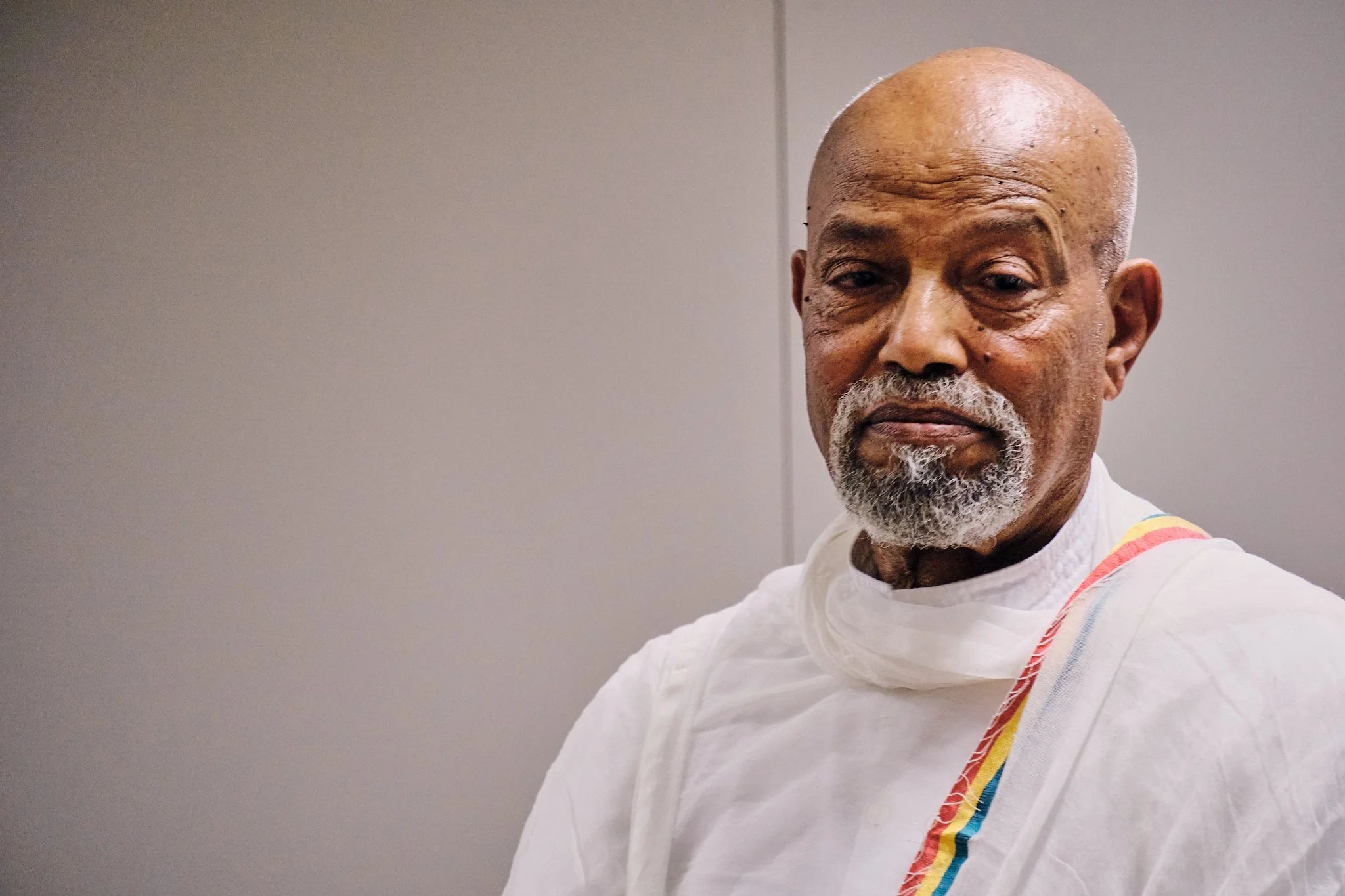 Close-up of an elderly man with a bald head and gray beard, wearing white traditional attire with colorful rainbow stripes on the shoulder, standing against a plain background.
