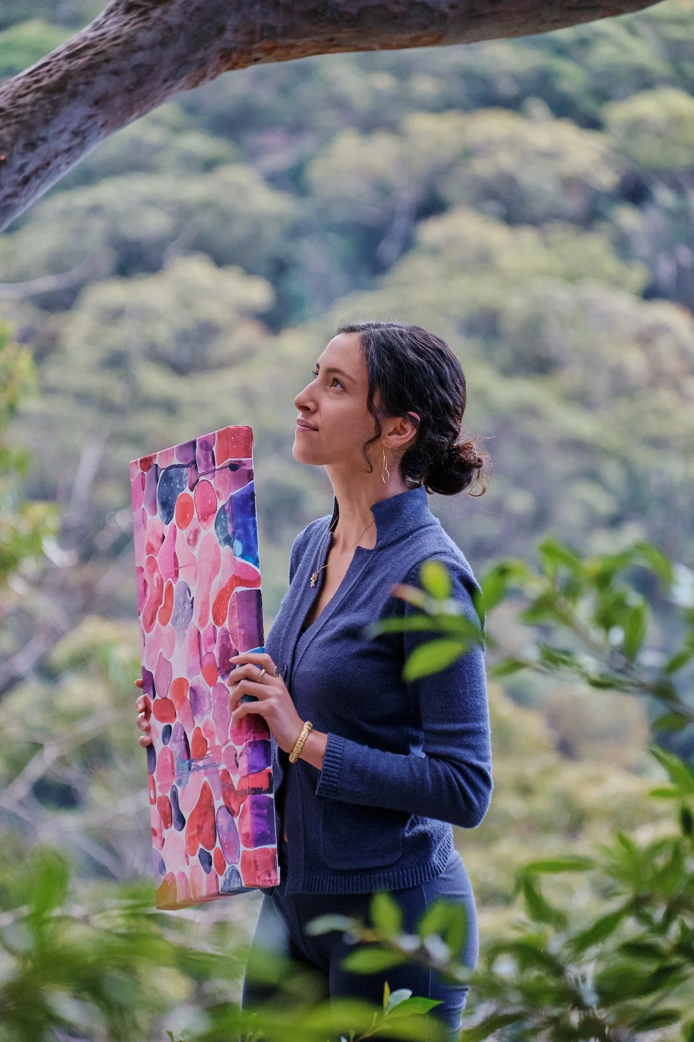 A woman standing outdoors in front of a blurred forest background, holding a colorful abstract painting with shades of pink, purple, and blue, and looking upwards.