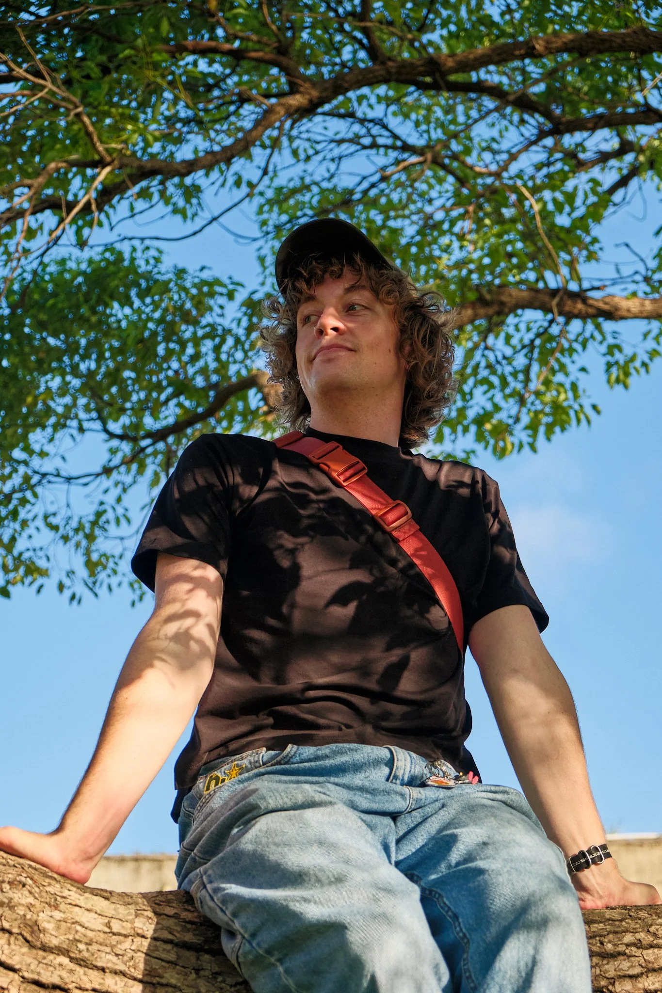 A young man sitting on a tree branch, looking to the side with a slight smile. He is wearing a black t-shirt, faded jeans, a black cap, and a red strap across his chest. There are green leaves and a clear blue sky in the background.