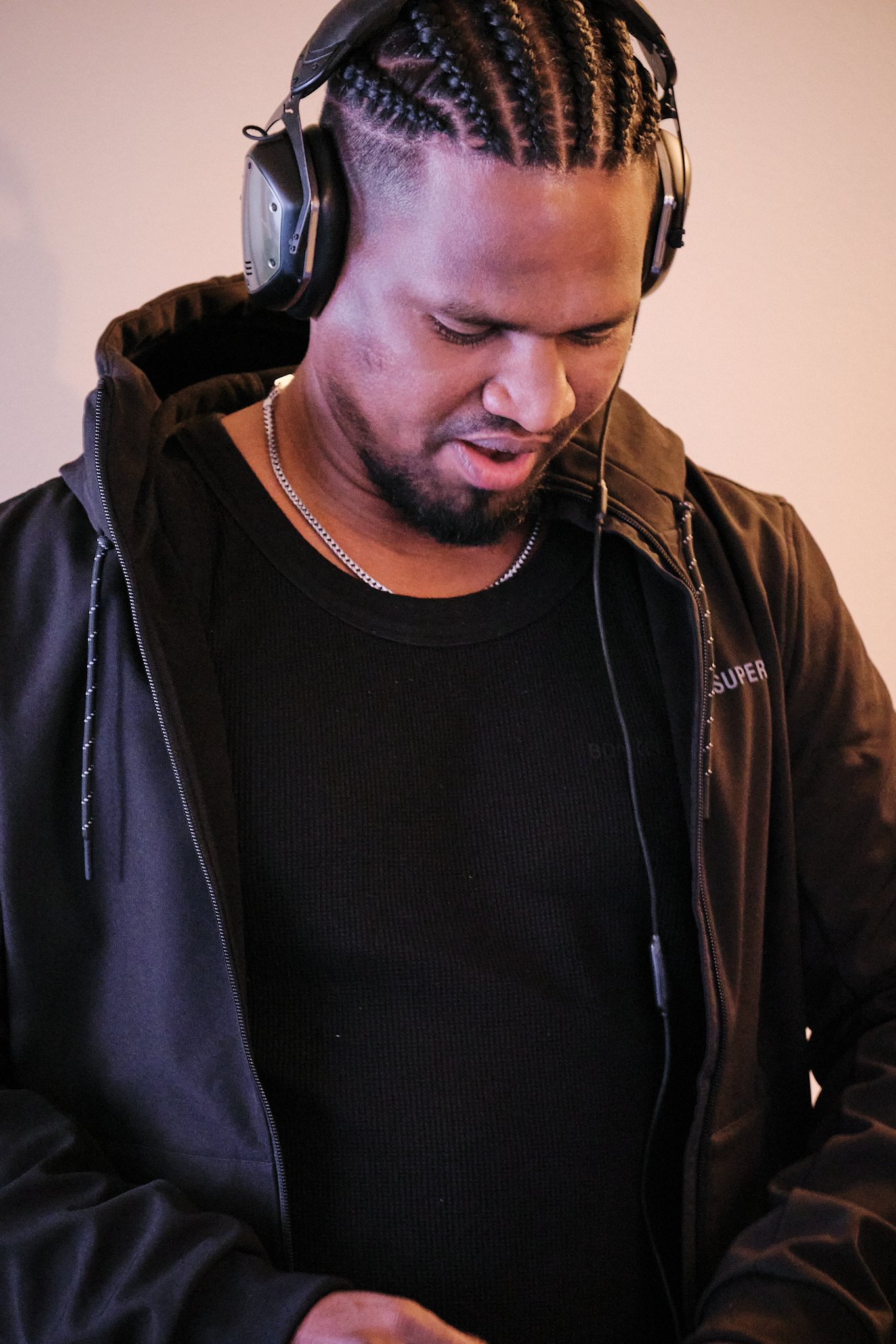 A man wearing headphones, a black jacket, and a black shirt, looking down with a slight smile.