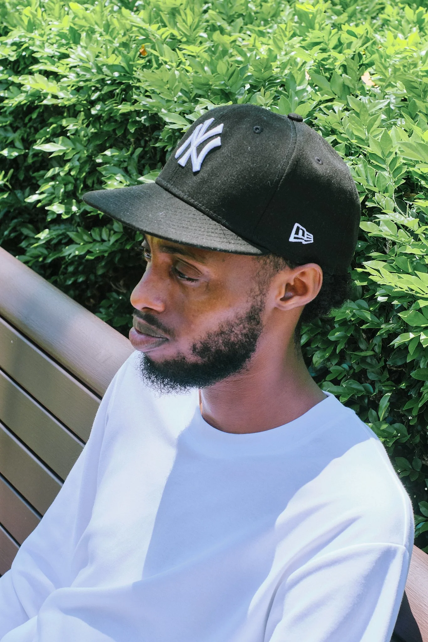 A young man in a white shirt and black New York Yankees cap with a white logo, sitting on a wooden bench with green foliage background.