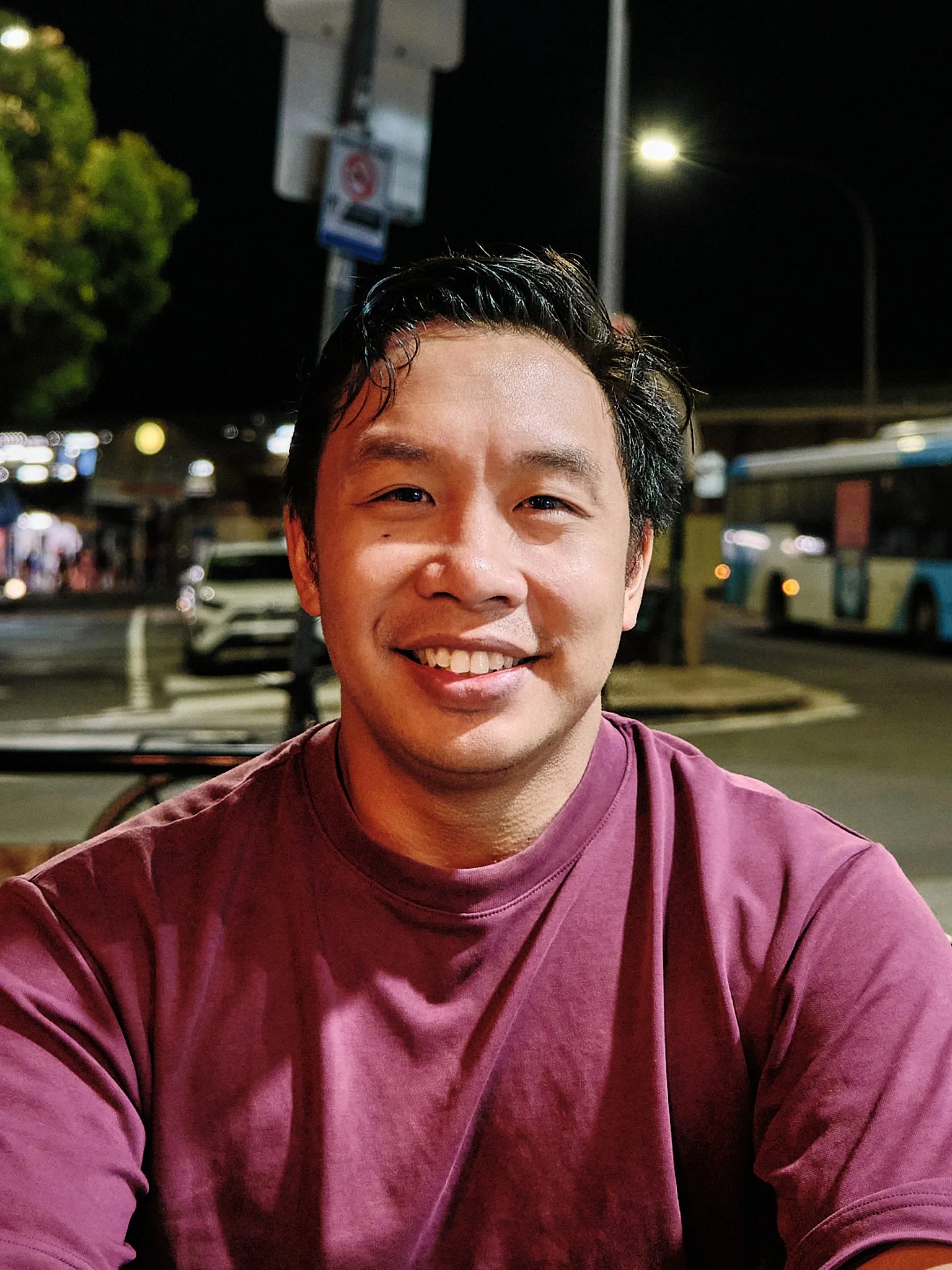 A smiling man wearing a maroon shirt sitting outdoors at night with streetlights and a bus in the background.