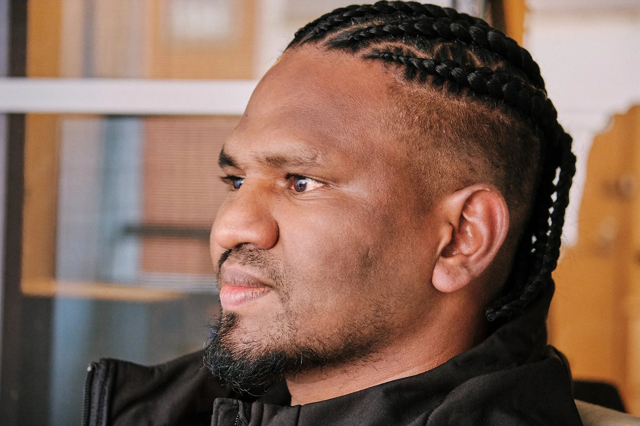 Close-up side profile of a man with braided hairstyle and beard, wearing a black jacket.