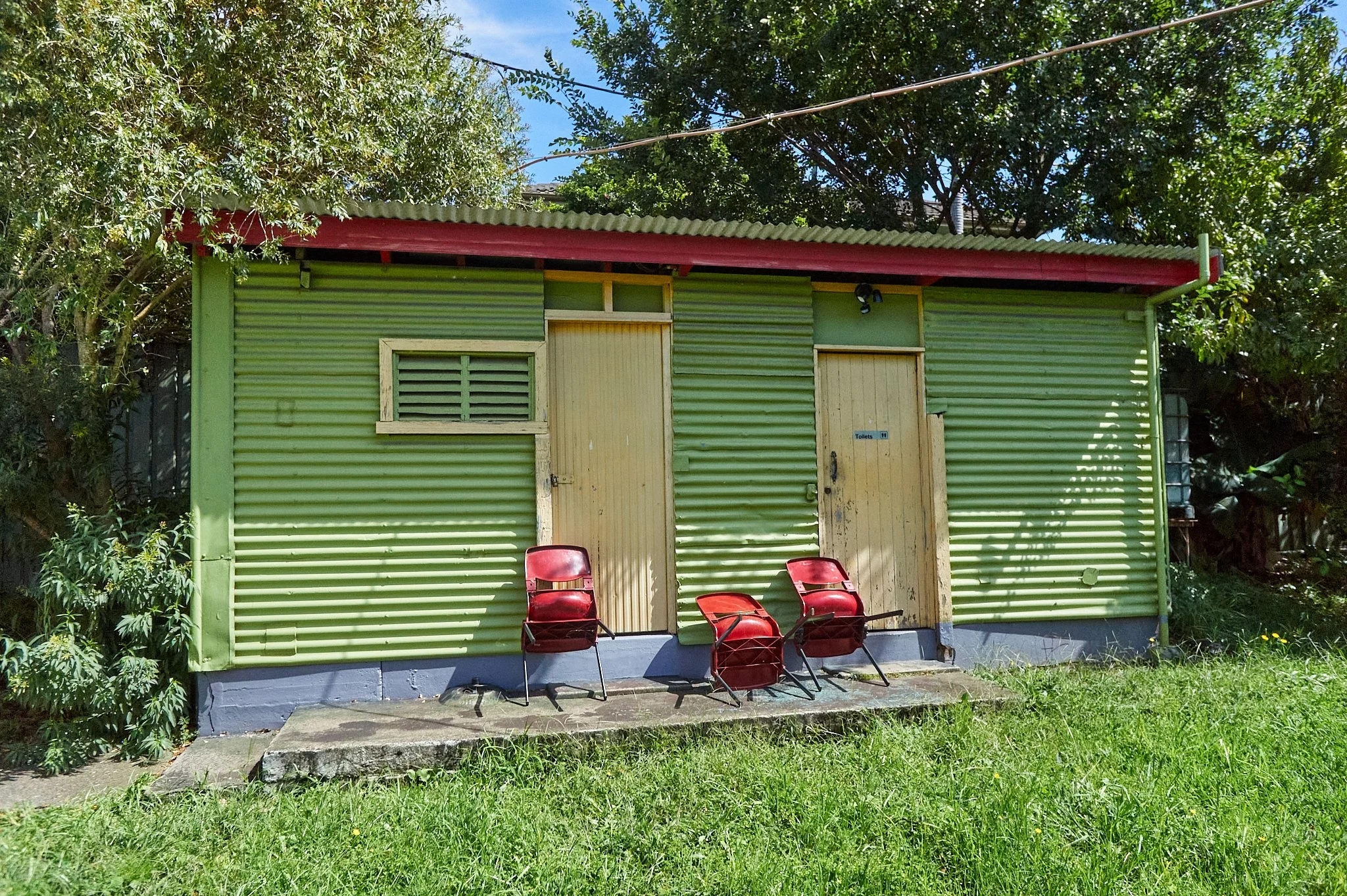 Small green corrugated metal building with two doors, one with a sign indicating toilets, three red chairs in front, grass and trees surrounding the building.