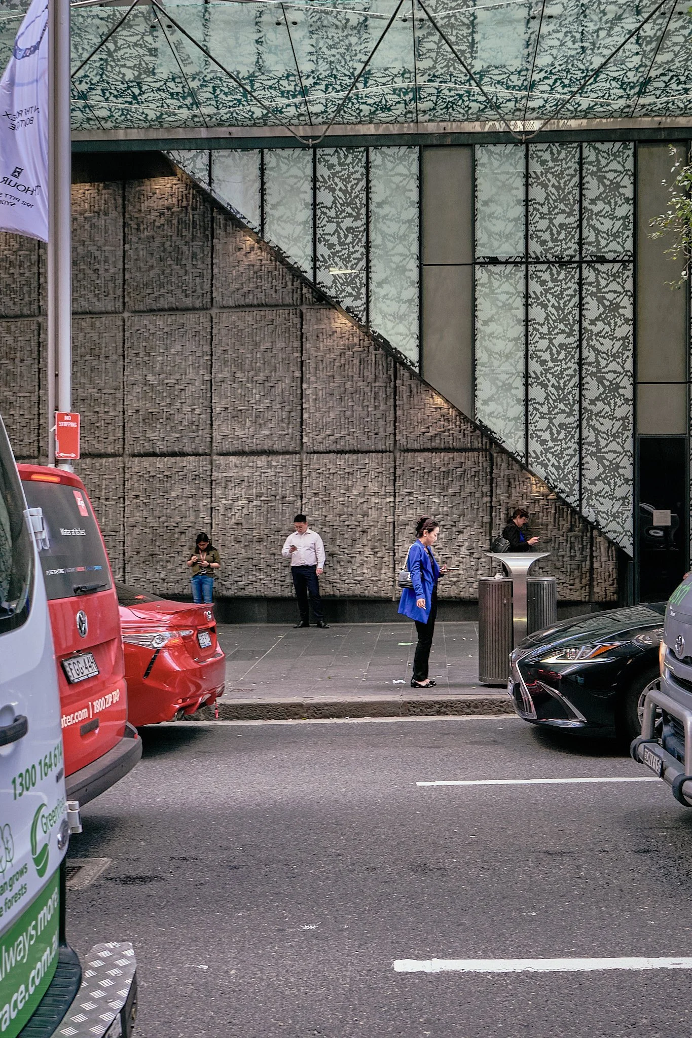 People standing and using their phones on a city sidewalk near parked cars, with a modern building featuring textured and patterned glass panels in the background.