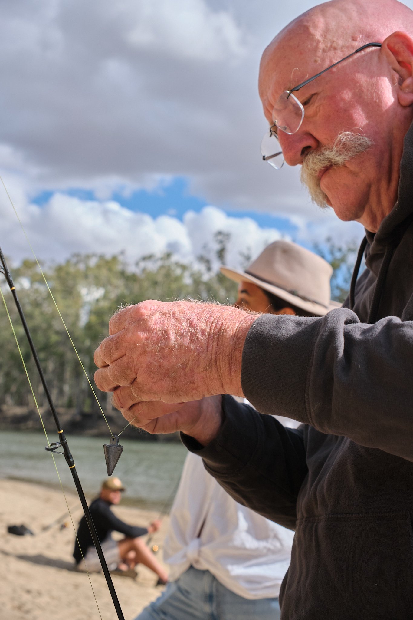 An older man with glasses and a mustache fishing on a beach under a partly cloudy sky, with a woman sitting in the background wearing a hat.