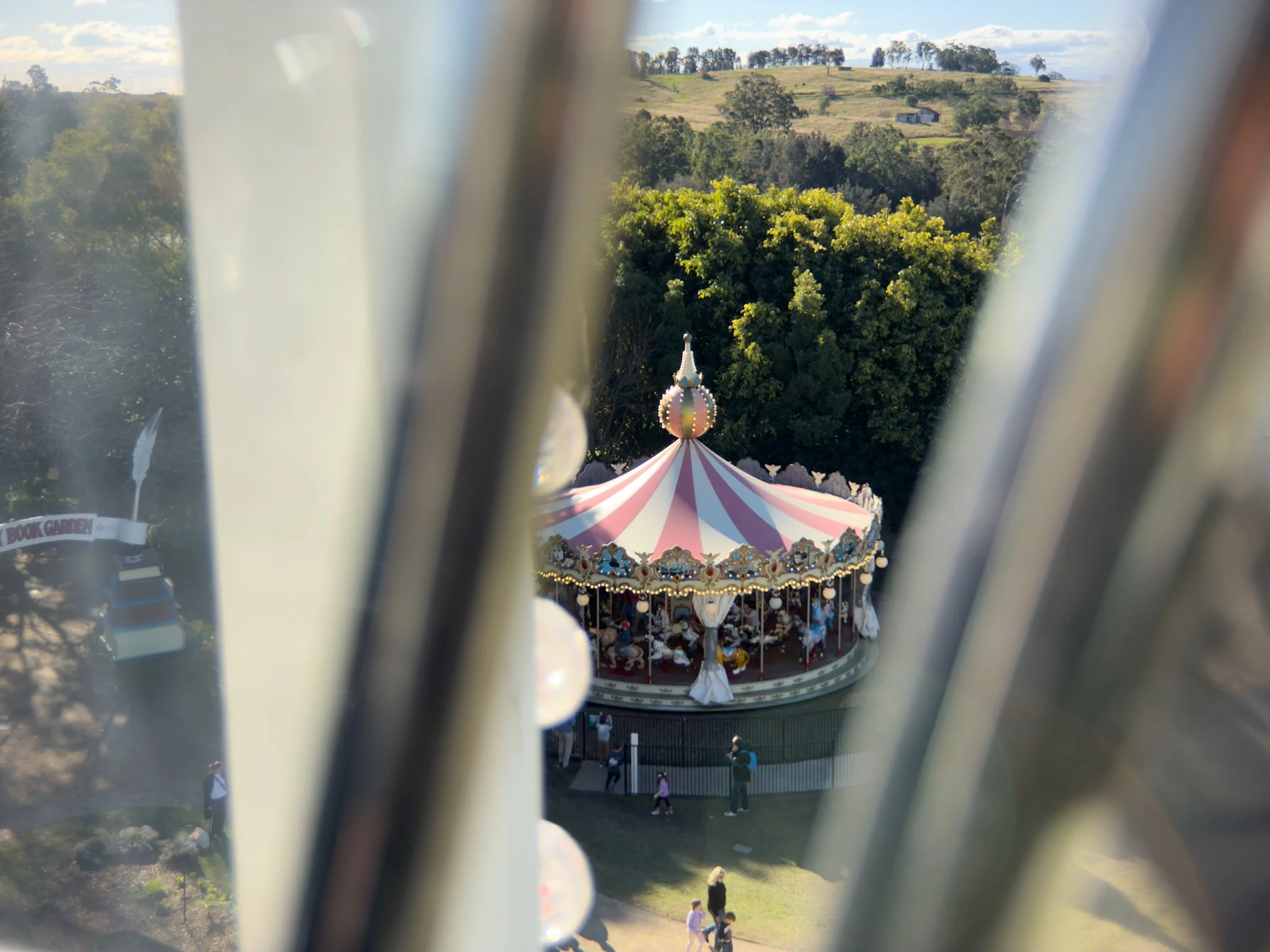 Photo taken from a hot air balloon, showing a carousel with a pink and white striped canopy in a park with trees and rolling hills in the background.