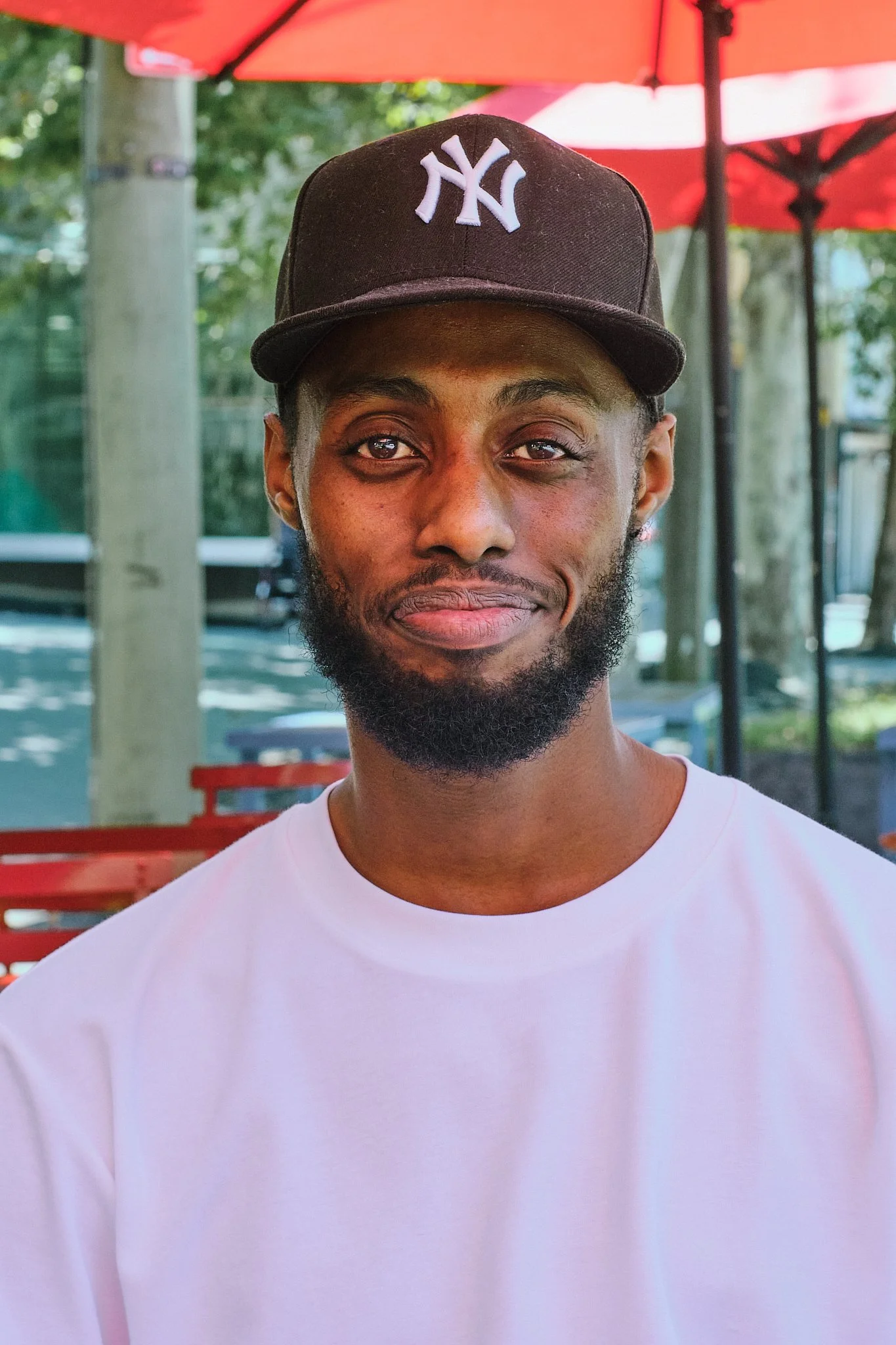A man with a beard wearing a black New York Yankees baseball cap and a white T-shirt, sitting outdoors under a red umbrella with trees and yard furniture in the background.