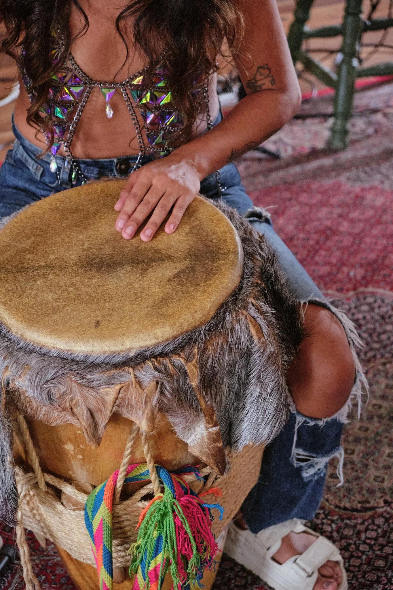 Person with long hair and tattoos playing a hand drum, decorated with colorful woven straps, on a patterned carpet.