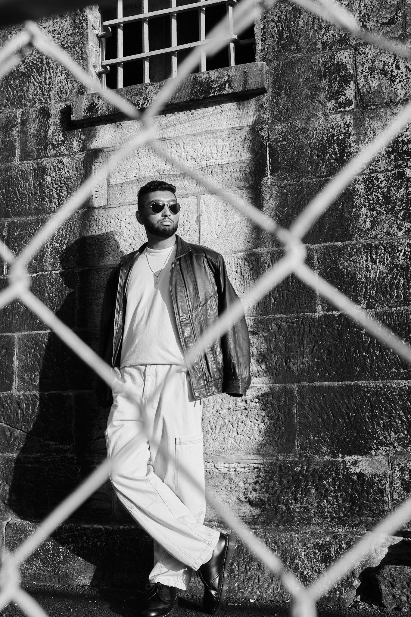 A man wearing sunglasses, a leather jacket, and white pants, leaning against a stone wall with a barred window above him, viewed through a chain-link fence, in a black and white photo.