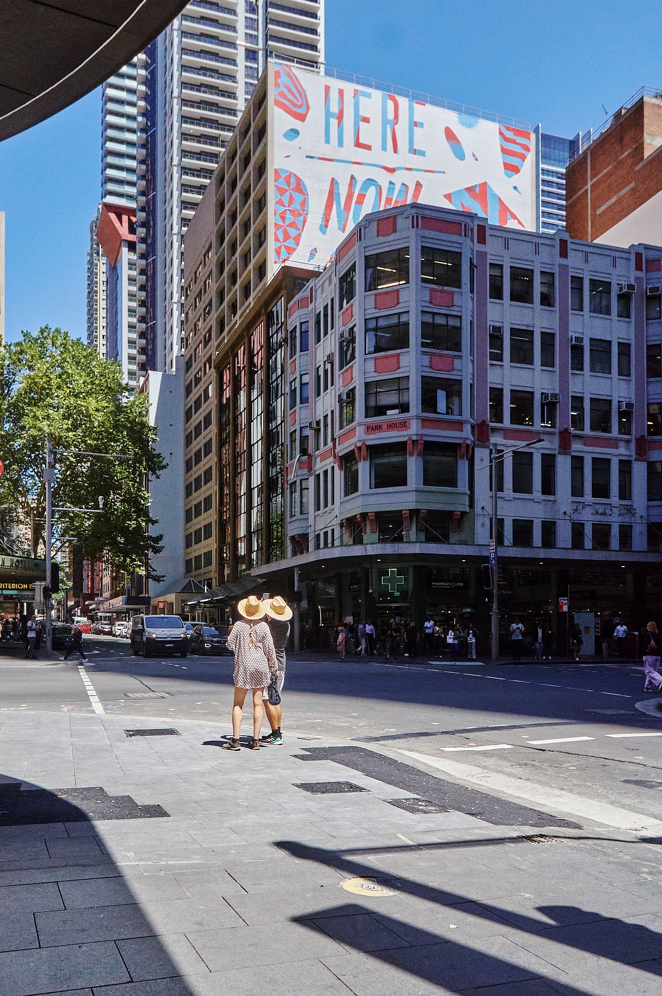 City street scene with tall buildings, pedestrians, and a digital billboard that says "HERE NOW." Two people wearing large wide-brimmed hats are crossing the intersection, while other pedestrians wait on the sidewalk. There are cars on the street and