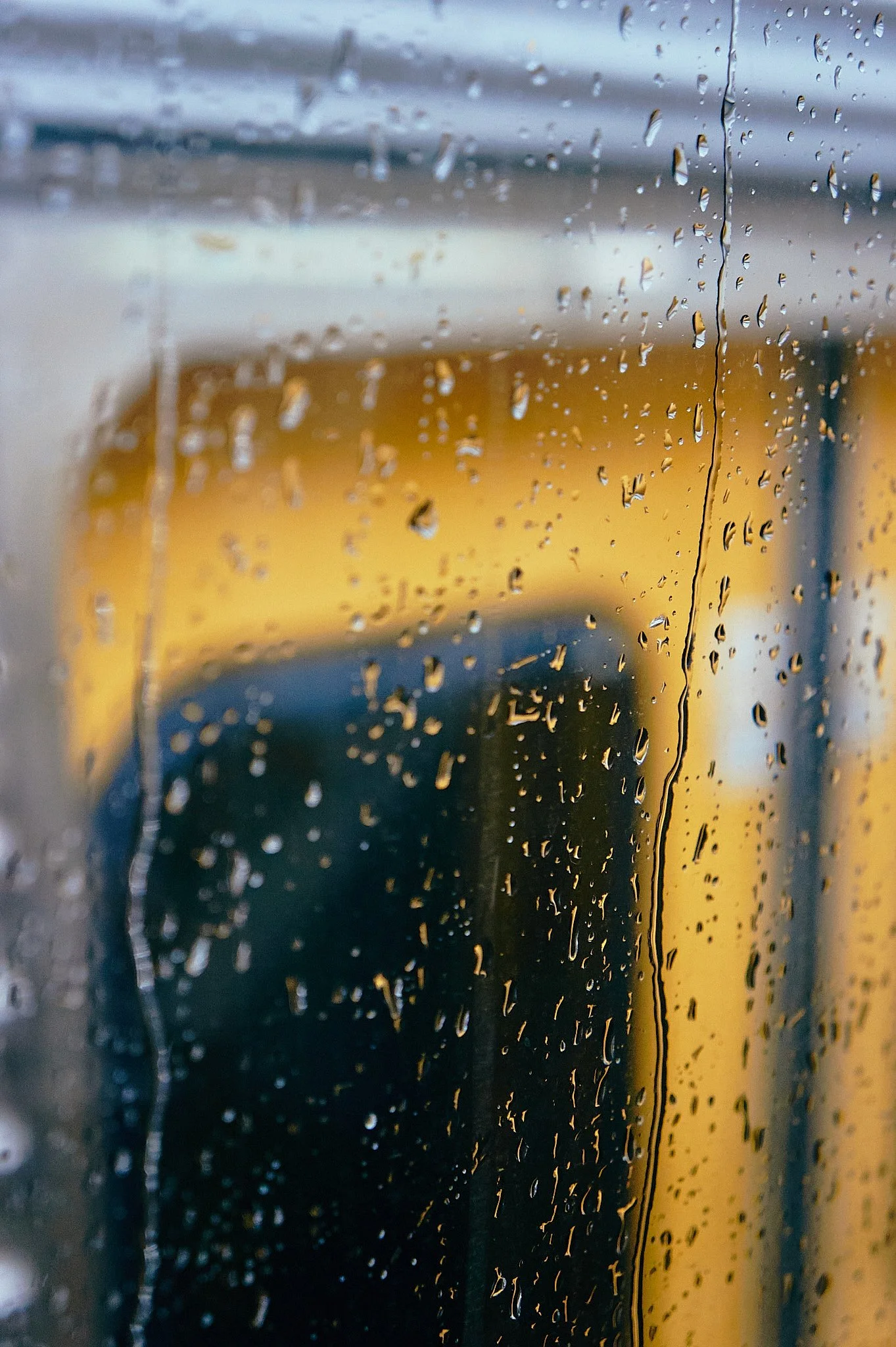 Rain droplets on a glass window with blurred yellow and dark background behind it.