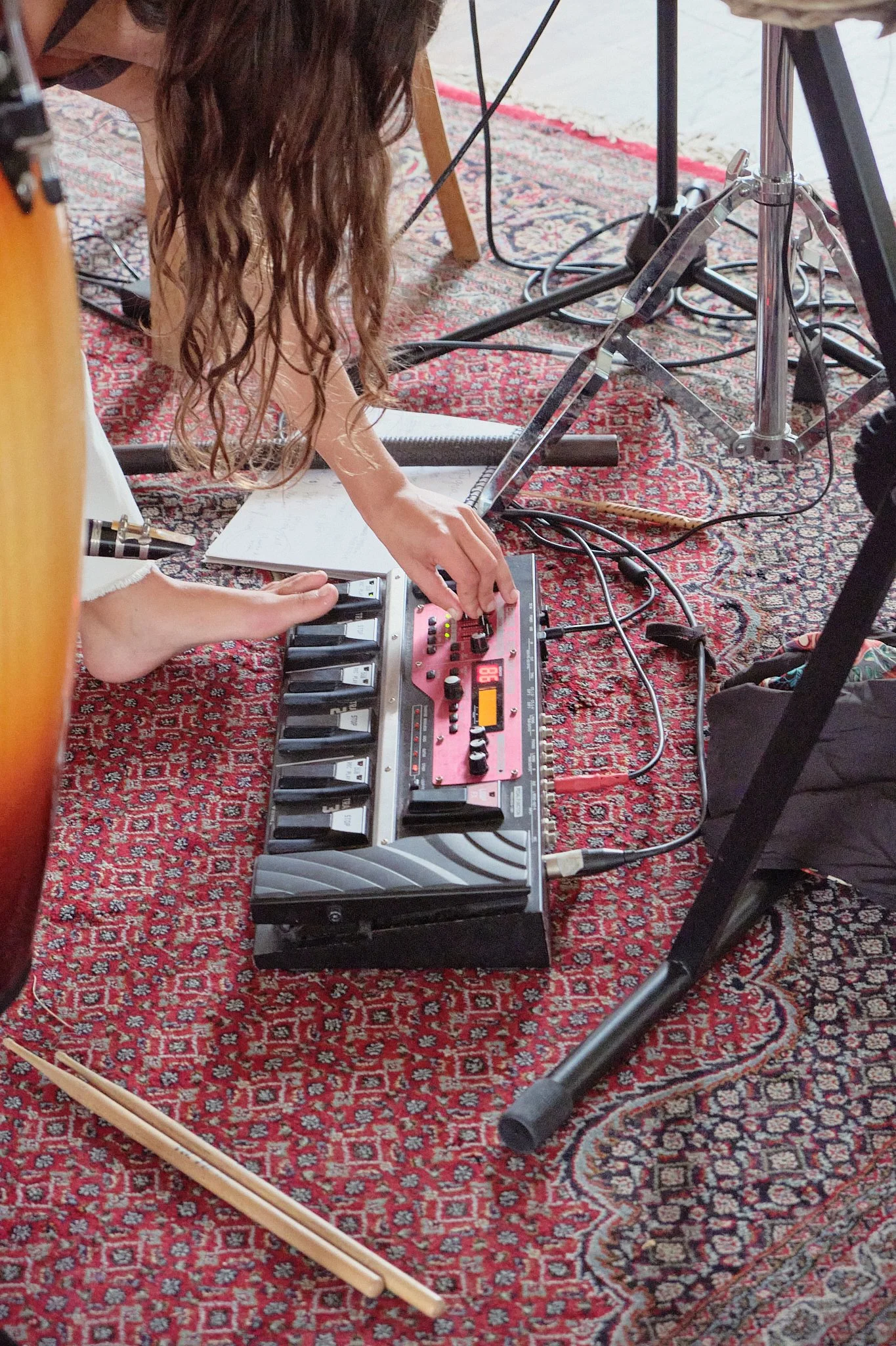 Person adjusting electronic music equipment on a patterned rug, with musical instruments and stands visible in the background.