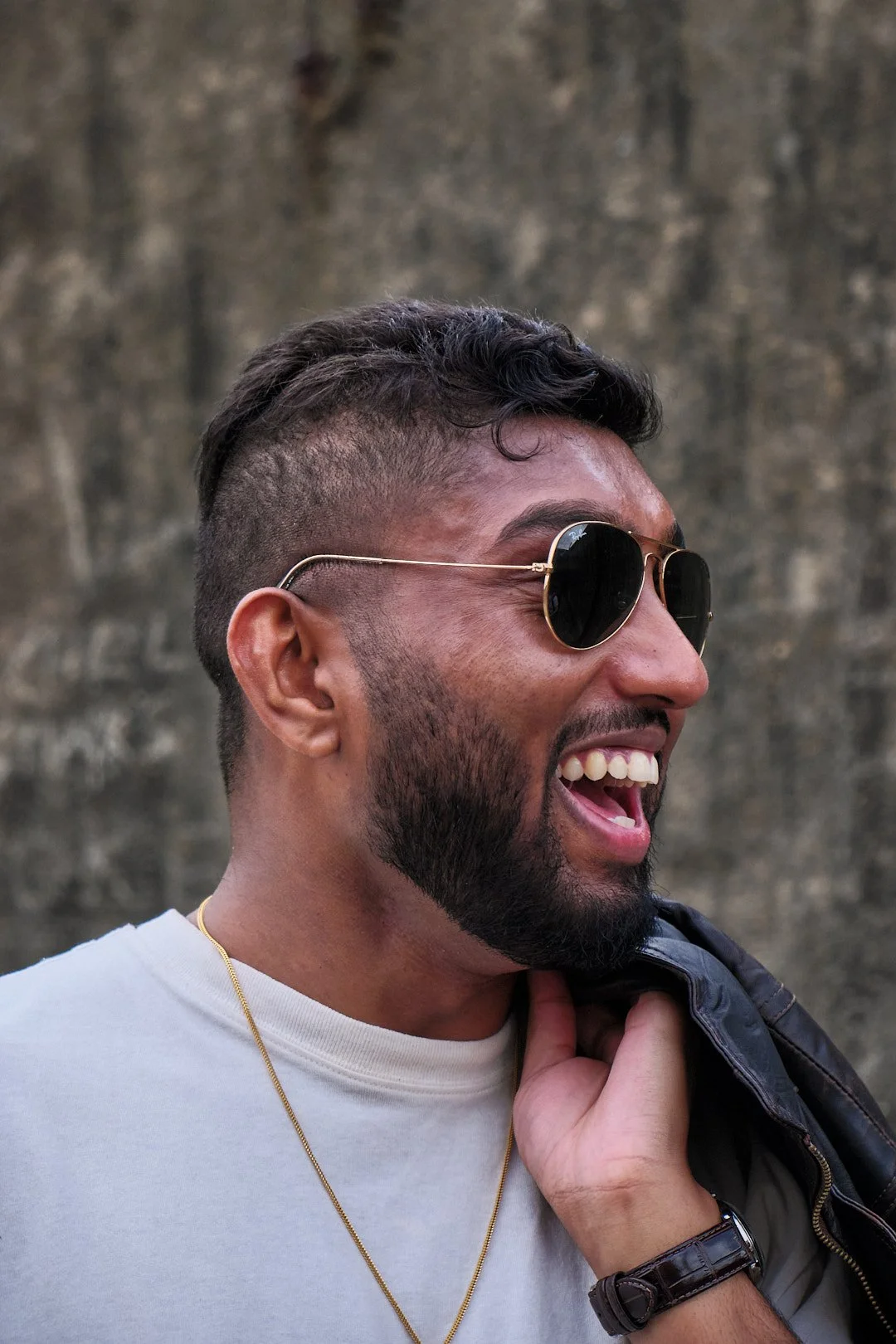 A man with a beard and sunglasses smiling and looking to the right, wearing a white shirt, a gold necklace, and a dark jacket over his shoulder, standing outdoors in front of a blurred stone wall.
