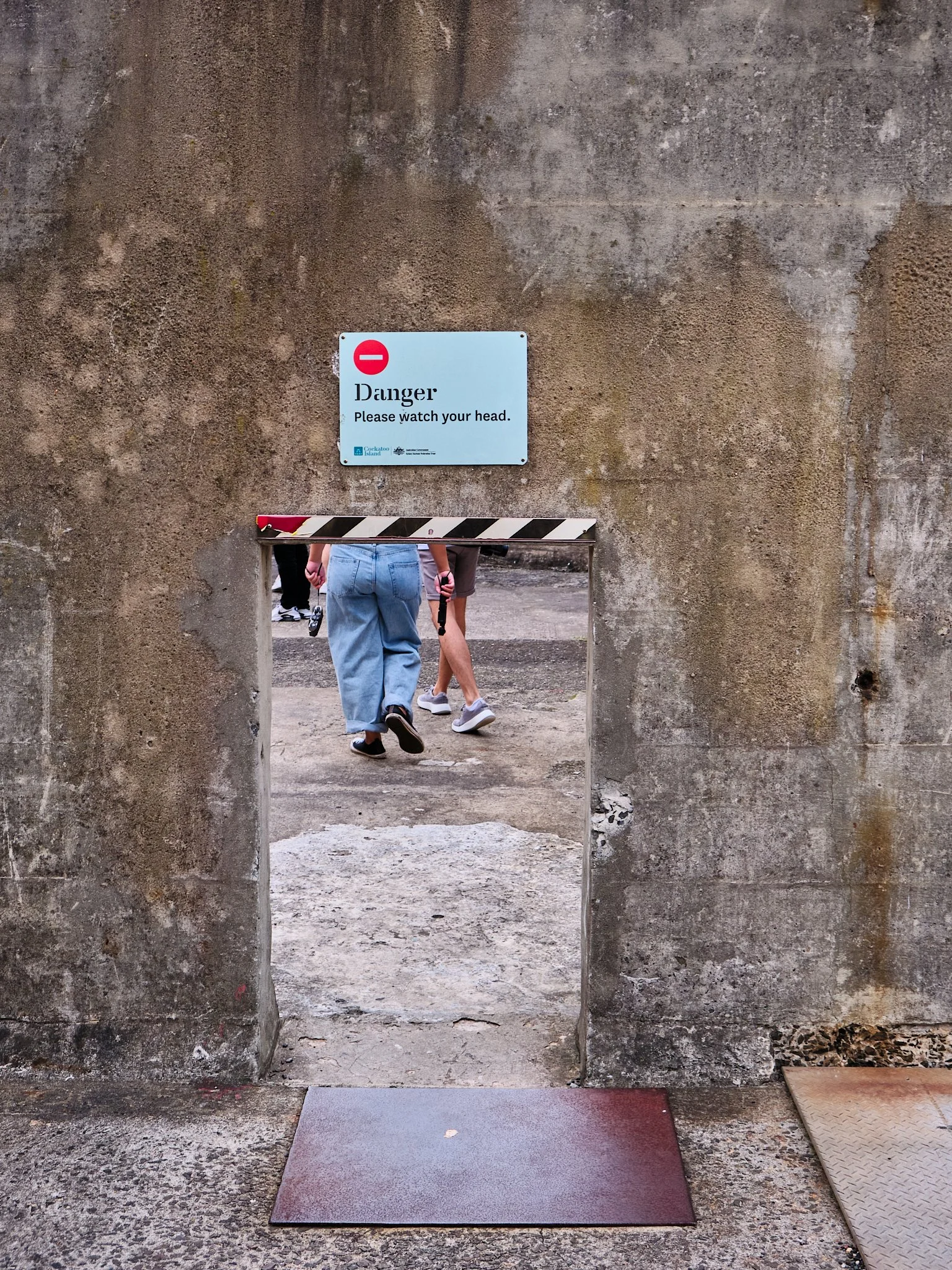 A small opening in a concrete wall with a warning sign above it that says 'Danger: Please watch your head.' Below the sign, a group of people are walking through, and their legs and feet are visible.