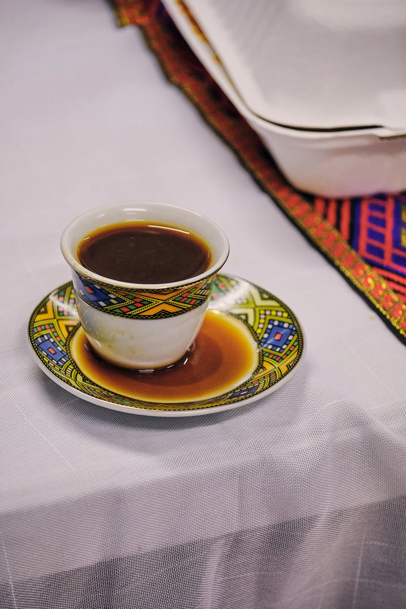 A traditional brewed Ethiopian Coffee drink served in a traditional Ethiopian cup (Sini), decorated cup on a matching saucer resting on a white tablecloth, with a decorative cloth and a tray of papers in the background.