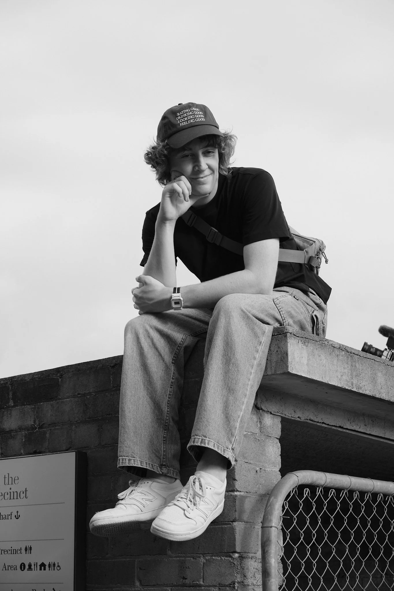 A young person with curly hair, wearing a cap with text, a black T-shirt, jeans, and white sneakers, sitting on a brick wall and smiling at the camera.