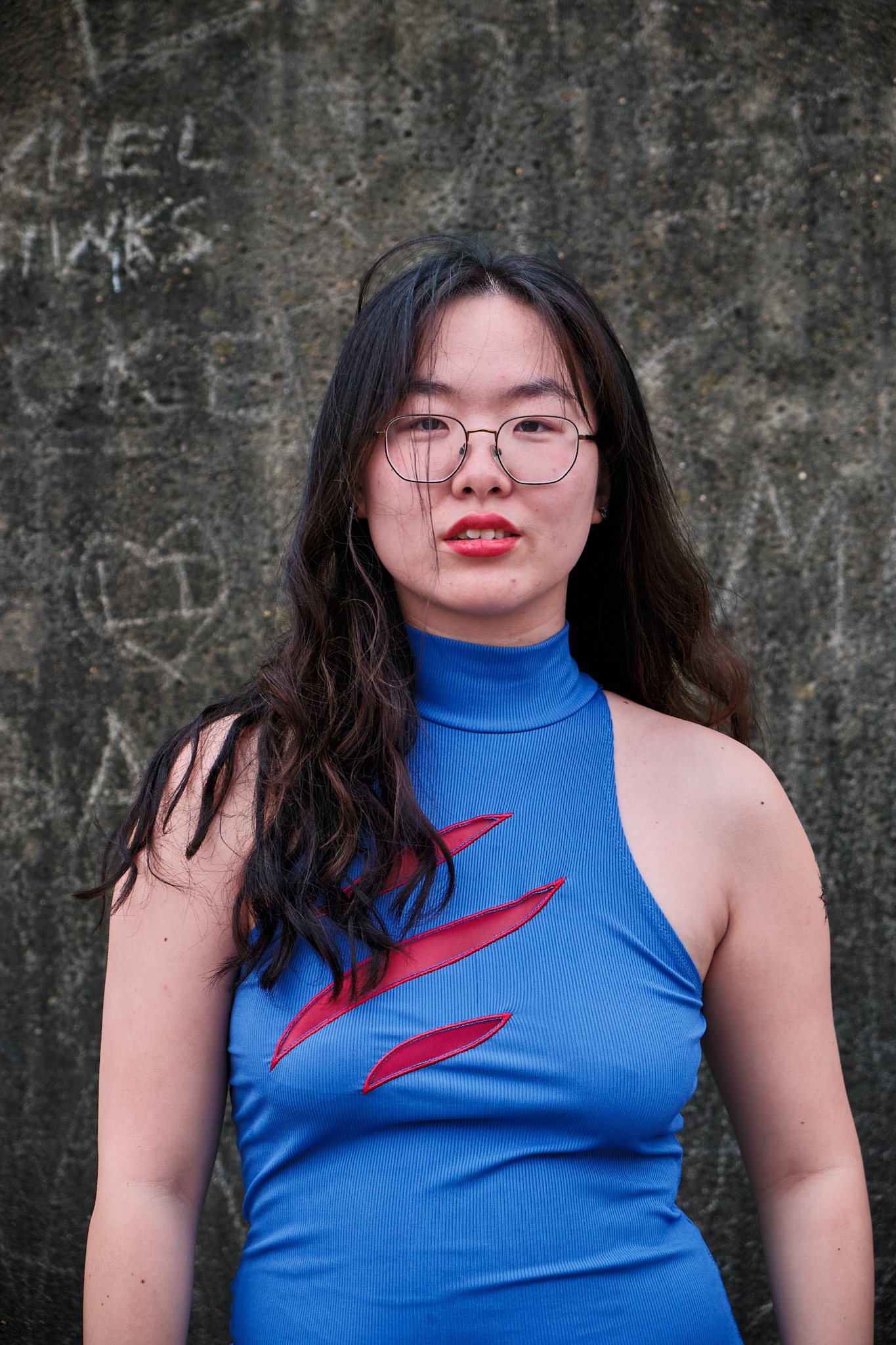 A young woman with long dark hair, wearing glasses and a blue sleeveless turtleneck top with red design, standing in front of a textured wall with faint graffiti or carvings.