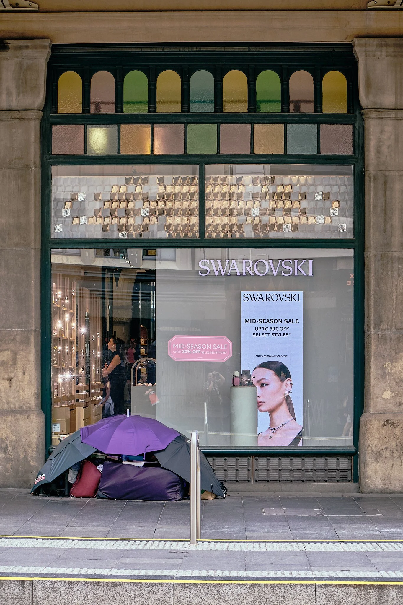 Store window for Swarovski with promotional signs about a mid-season sale, and items on display inside. There are some bags and jewelry inside, and a lone purple umbrella and belongings outside on the sidewalk.