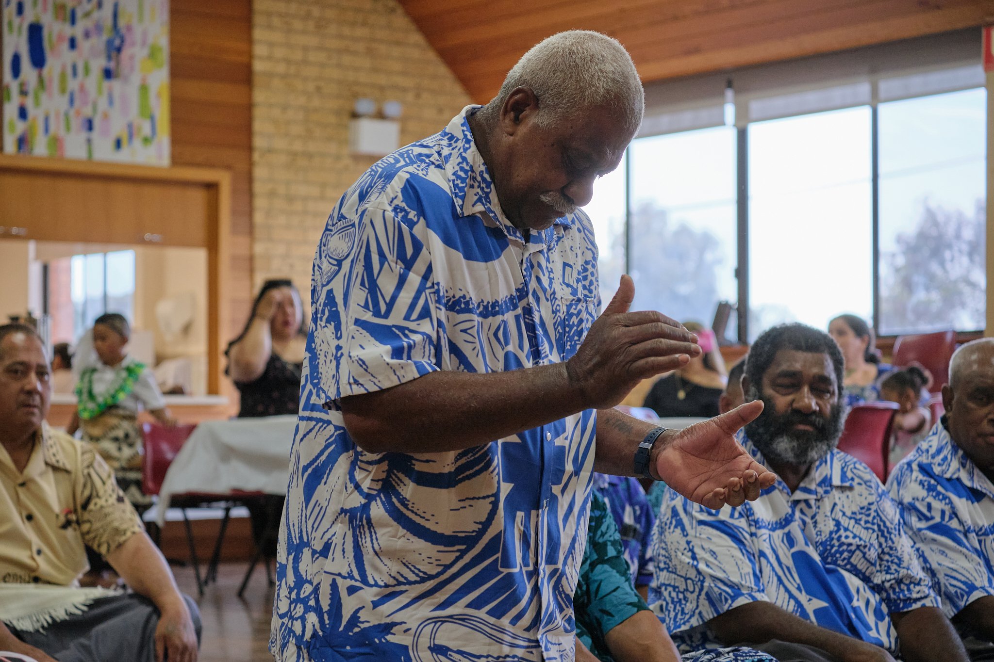 An elderly man with gray hair and a mustache, wearing a blue and white patterned shirt, is standing with his hands clasped as if praying or giving a blessing in a room with wooden walls and large windows. Several other people, some wearing matching p