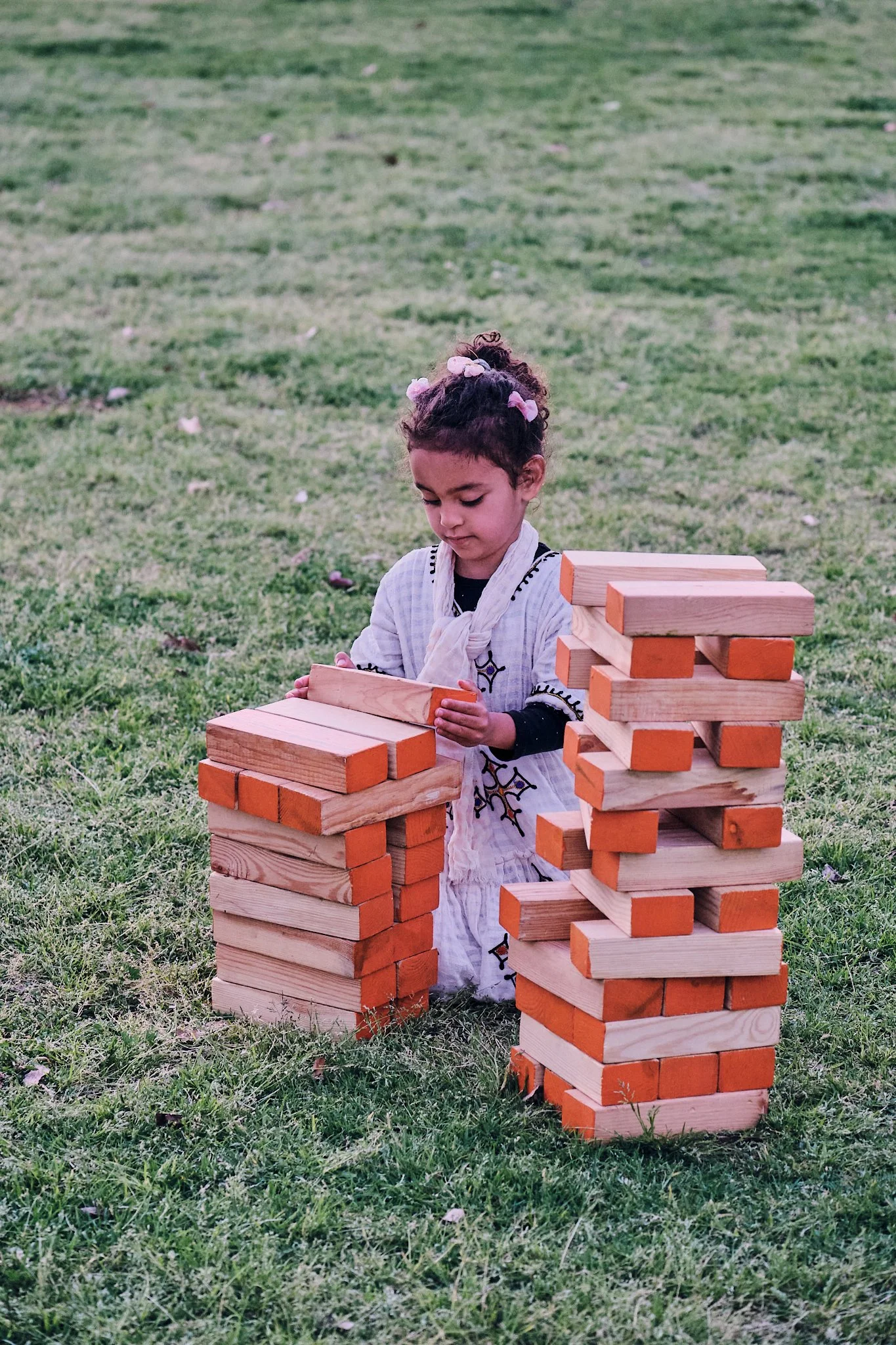 A young girl kneels on the grass, playing a giant game of Jenga with large wooden blocks.