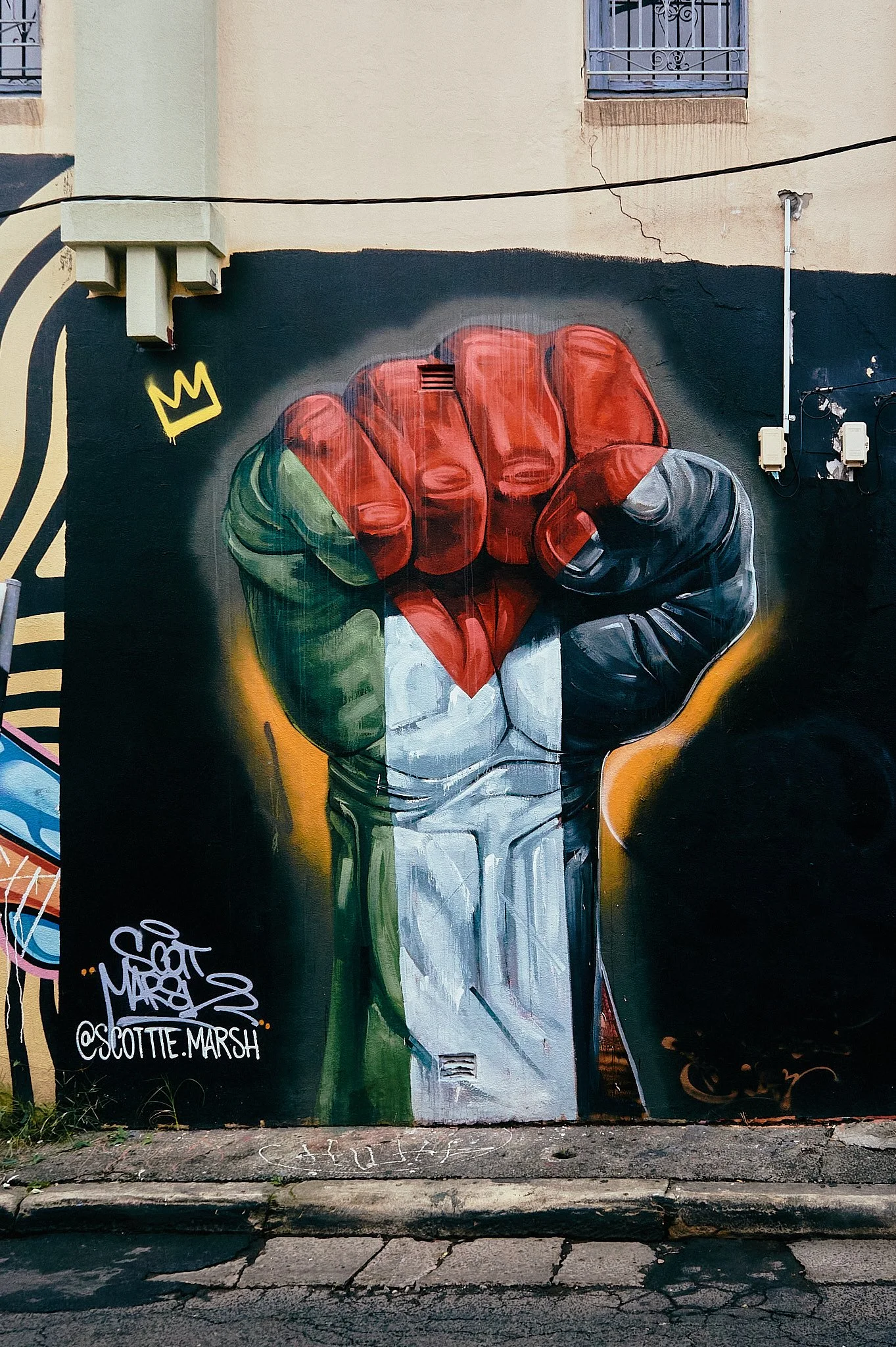 Street art mural of a clenched fist painted with the colors of the Italian flag, with a black background, a small yellow crown symbol, and artist's signature and social media handle at the bottom left.
