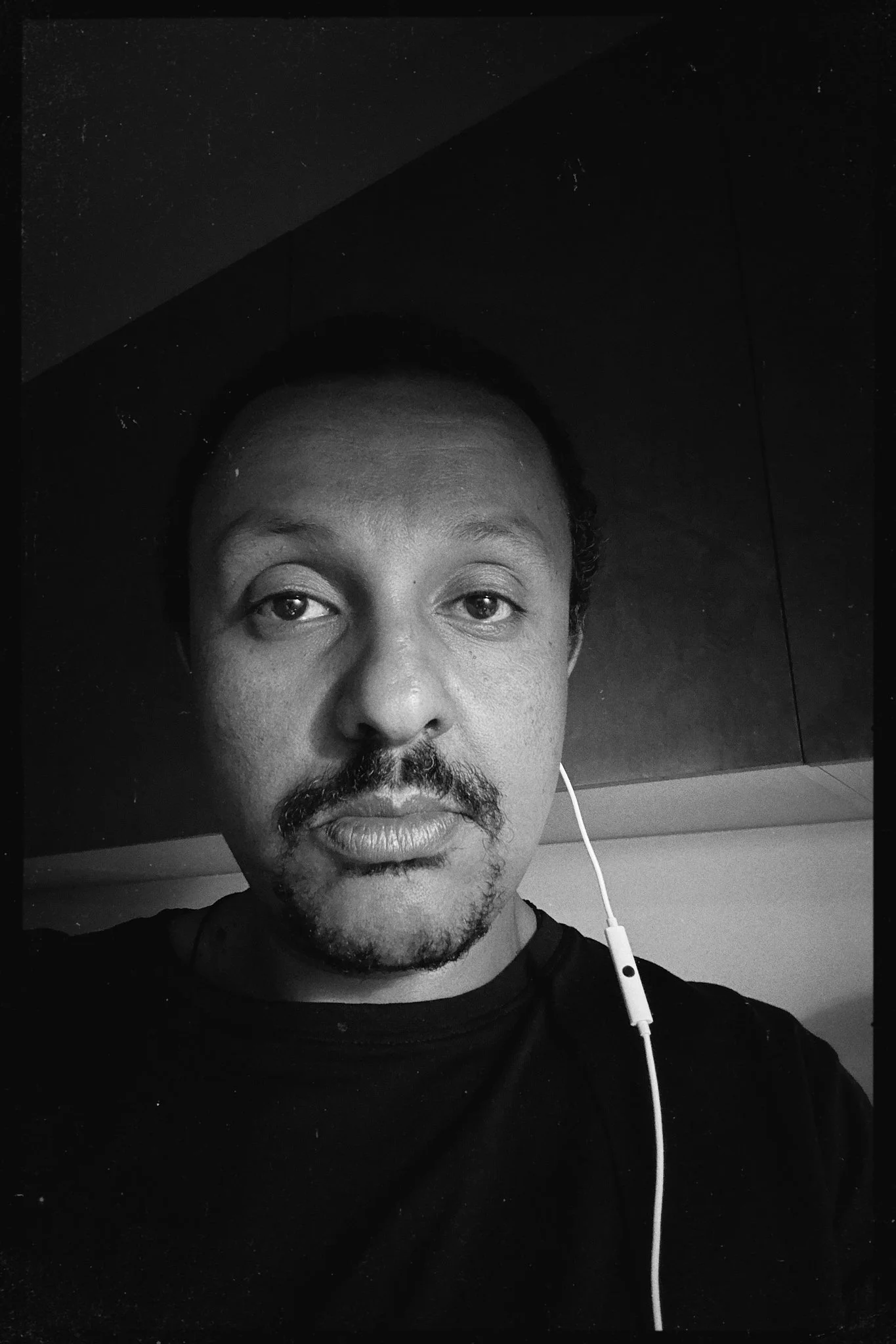 Black and white close-up selfie of a man with a mustache and goatee, wearing a black shirt and earphones, standing indoors against a dark wall.