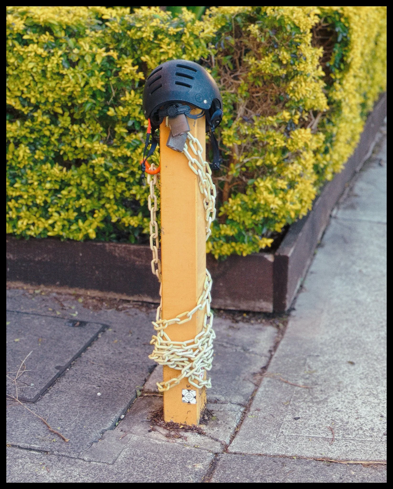 A black safety helmet placed on a yellow post wrapped with chains on a sidewalk with bushes in the background.