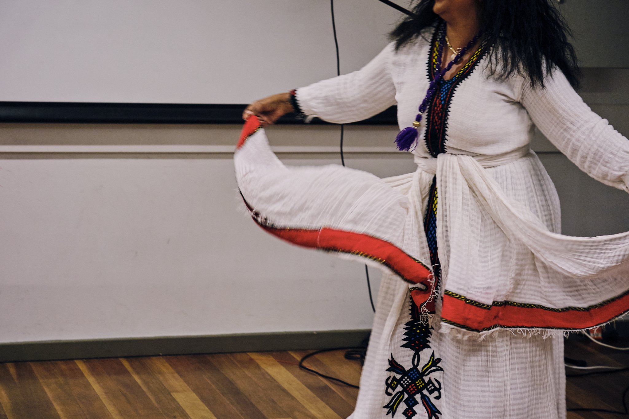 Woman in traditional embroidered white dress with red and blue accents, holding a flowing piece of fabric, possibly performing a traditional dance.