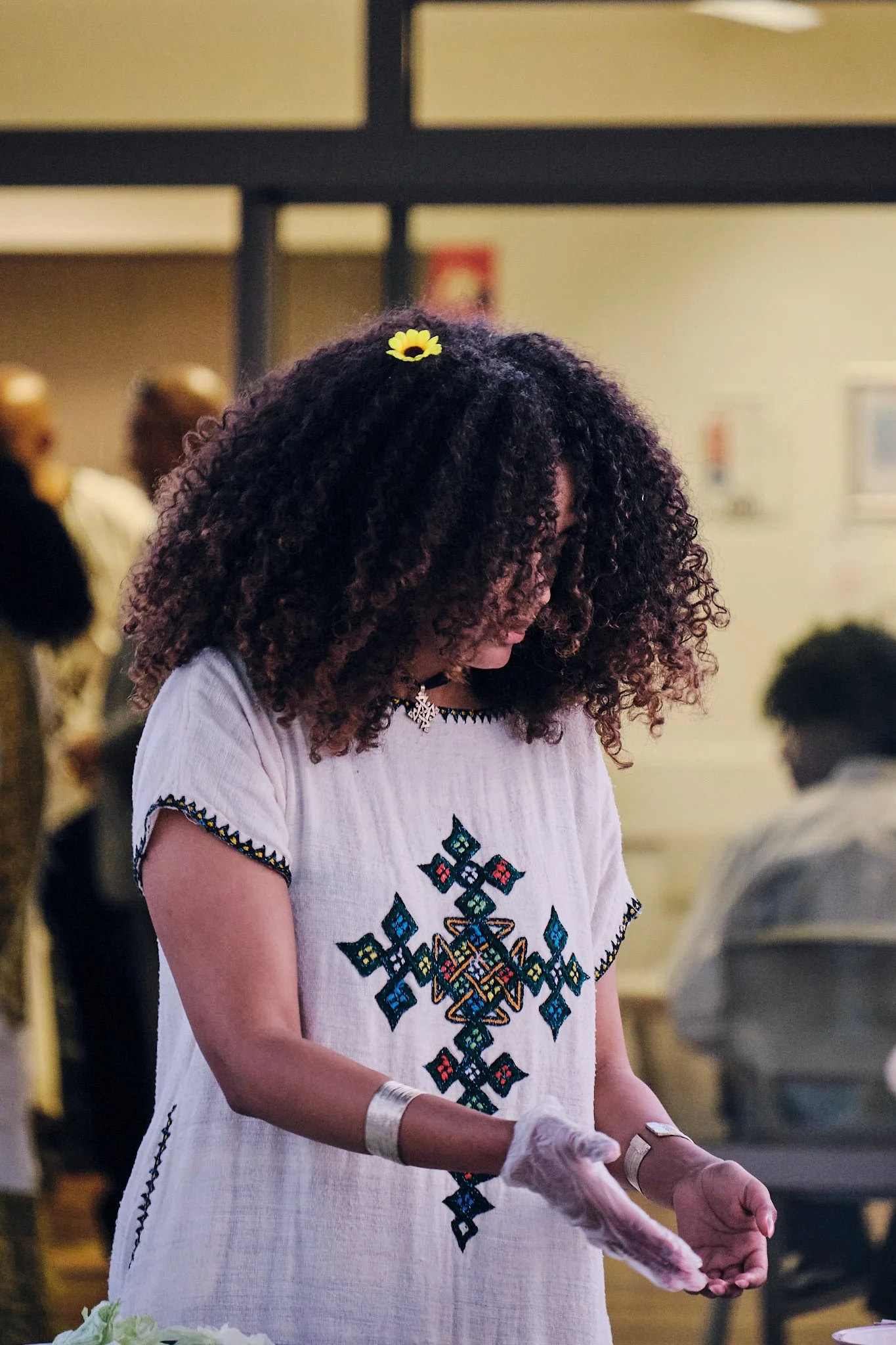 A woman with curly hair, wearing a white embroidered dress with blue and red patterns, is looking down and wearing gloves, at a social gathering.