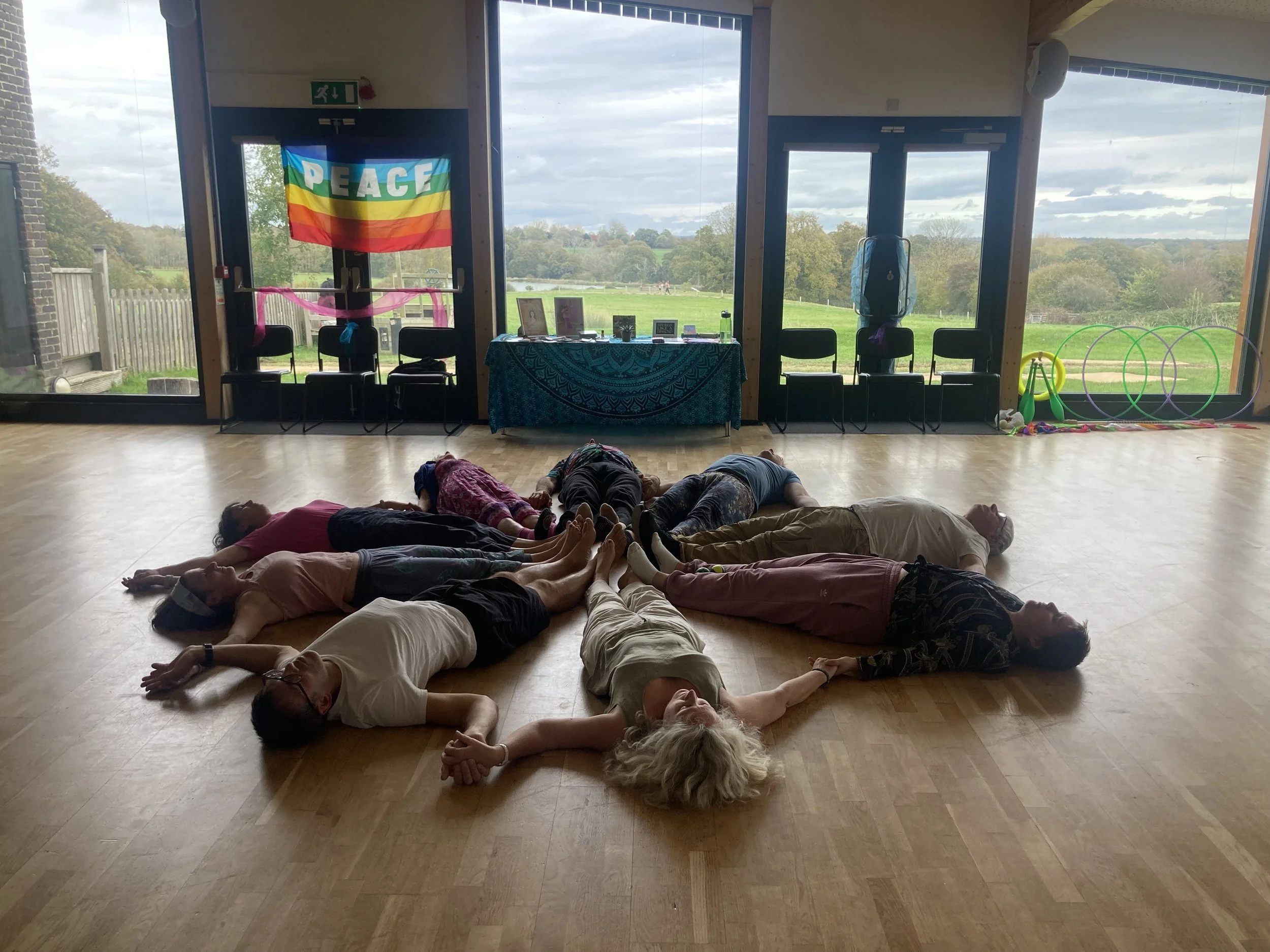 A group of people lying on the floor in a circle, holding hands, inside a spacious room with large windows overlooking a green landscape.