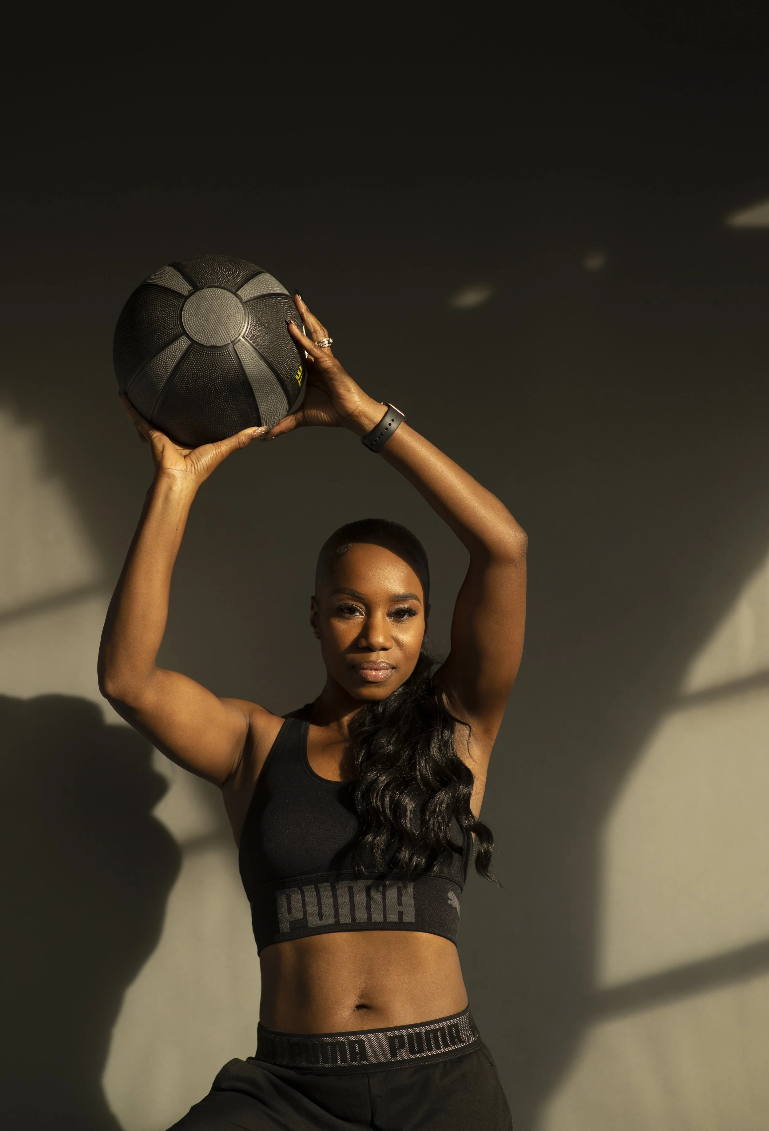 Vanessa Bosse une athlétique en vêtements de sport noirs tenant un ballon de basketball, debout devant un mur avec ombres et lumière naturelle.