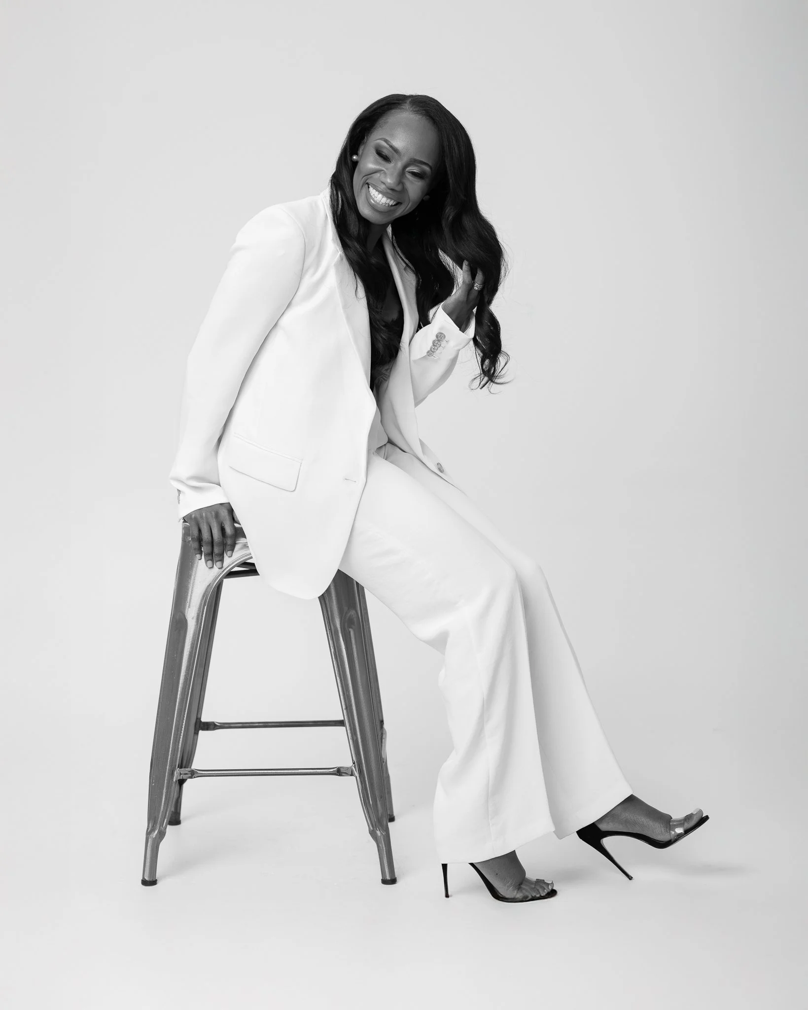 Une femme souriante, assise sur un tabouret, vêtue d'un tailleur blanc et de chaussures à talons noirs, en noir et blanc.