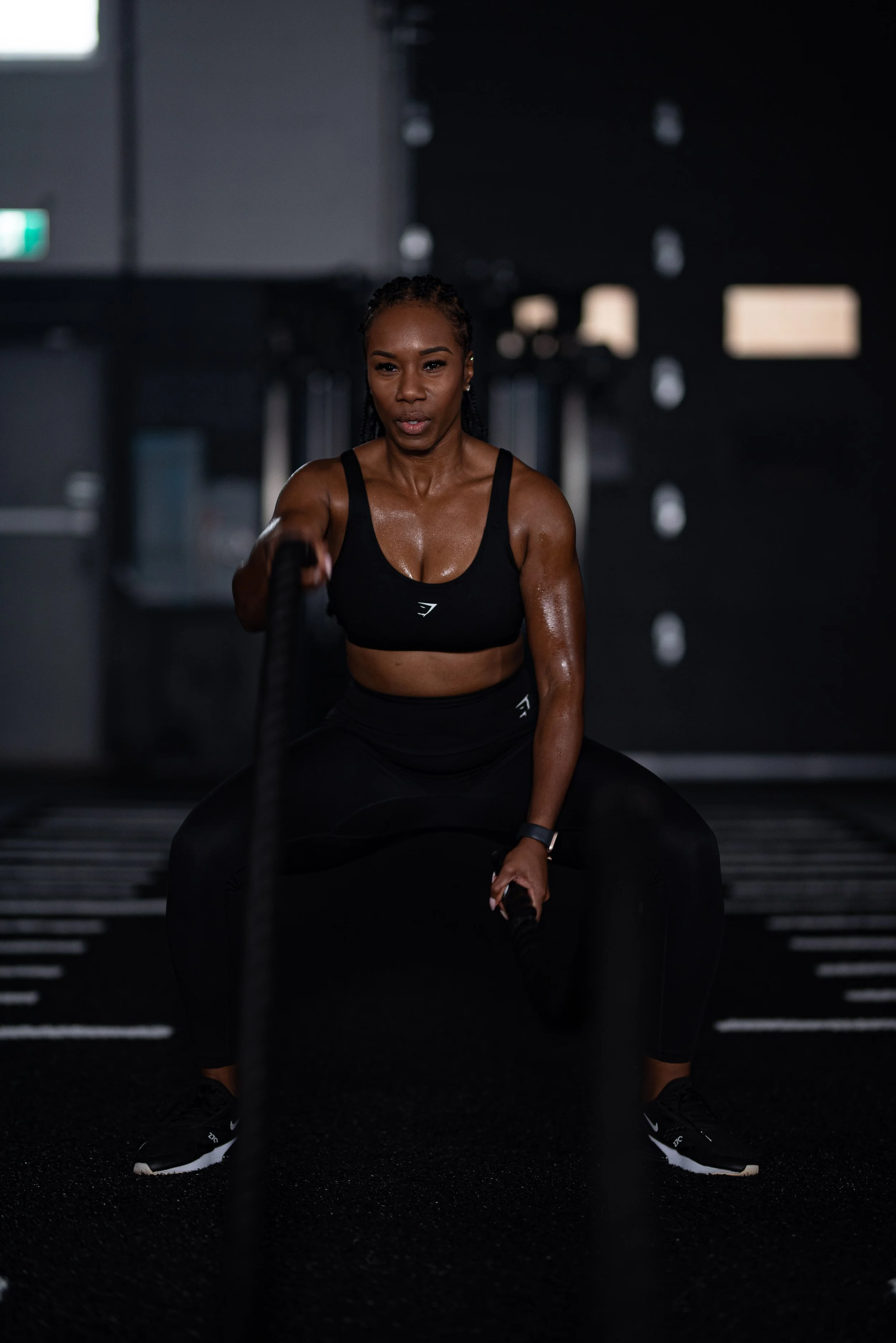 Vanessa Bosse musclée en tenue de sport effectue un exercice de musculation avec des chaînes dans une salle de sport sombre.