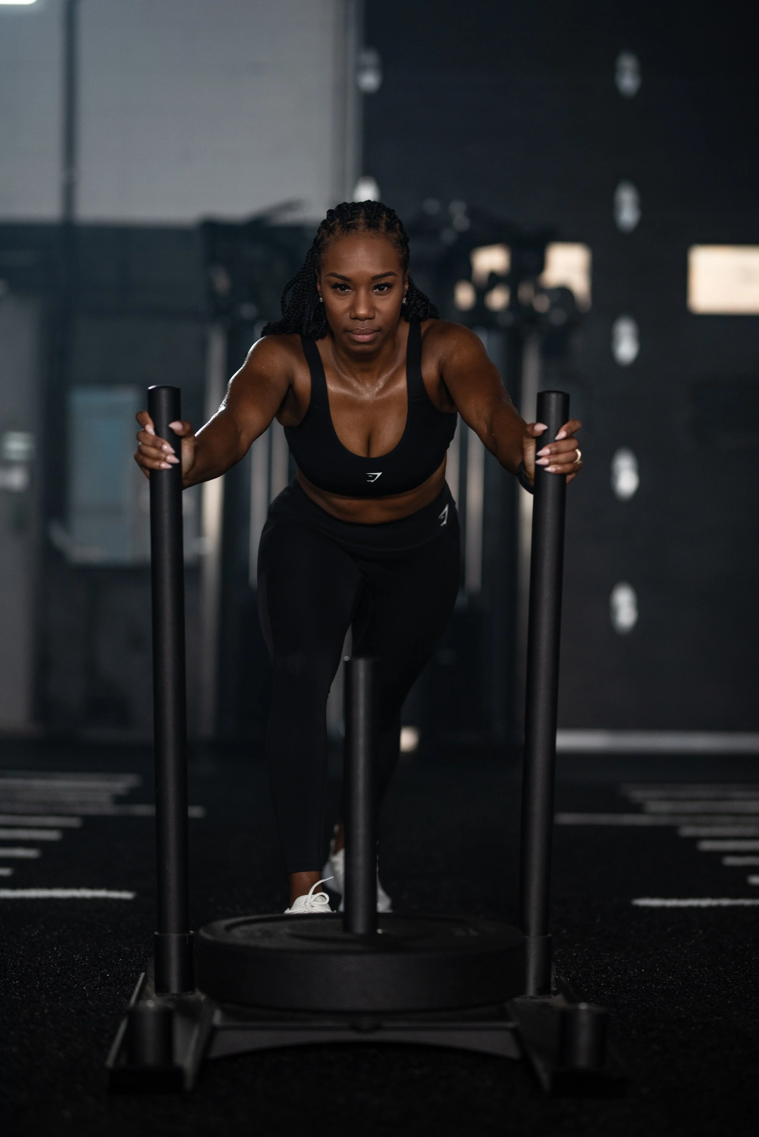 Vanessa Bosse faisante de l'exercice dans une salle de sport, utilisant un traîneau de force, avec un regard concentré et une tenue de sport noire.
