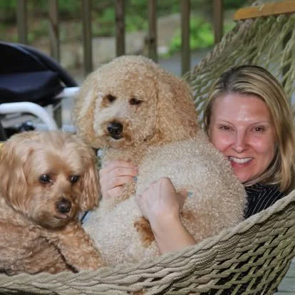 Jana smiling in a rope hammock, holding Tater, with Sassy perched beside them wearing a yellow flower.
