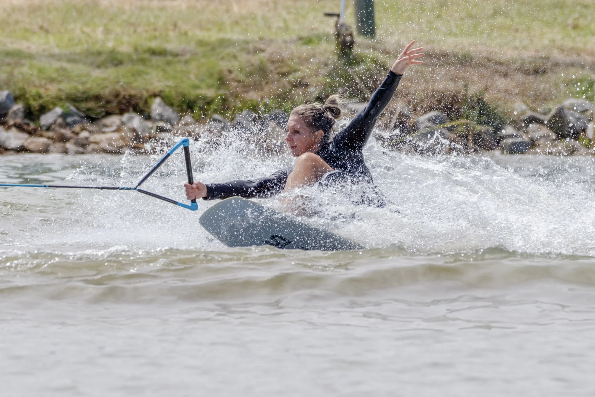 A woman (Jana Shelfer) on an adaptive trick sit ski carves hard through the water, one hand gripping the tow handle and the other arm raised, surrounded by spray.