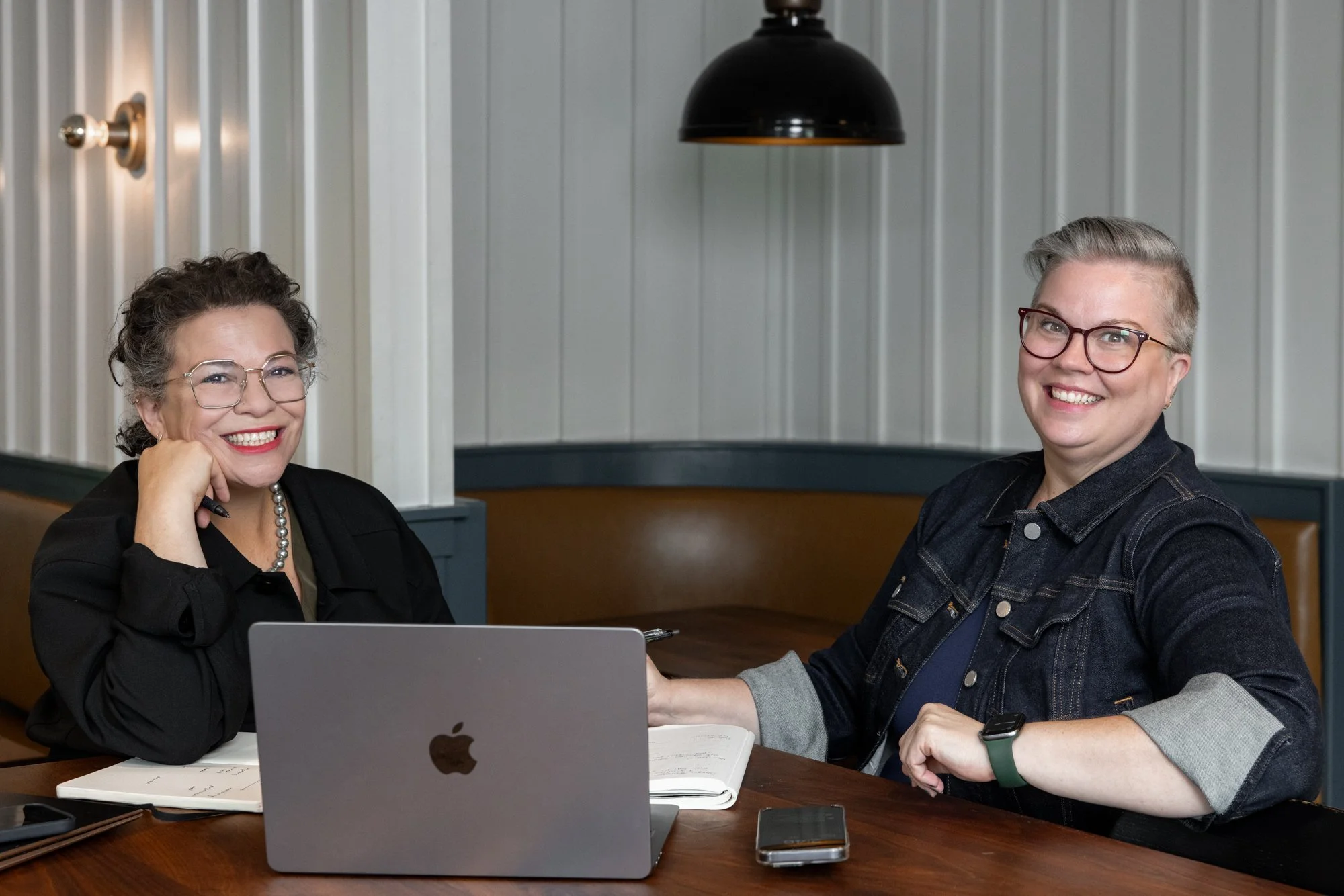 Lora Cover and Sarah Braver, leaders of coLeague, sit together at a table and are both smiling brightly at the camera.