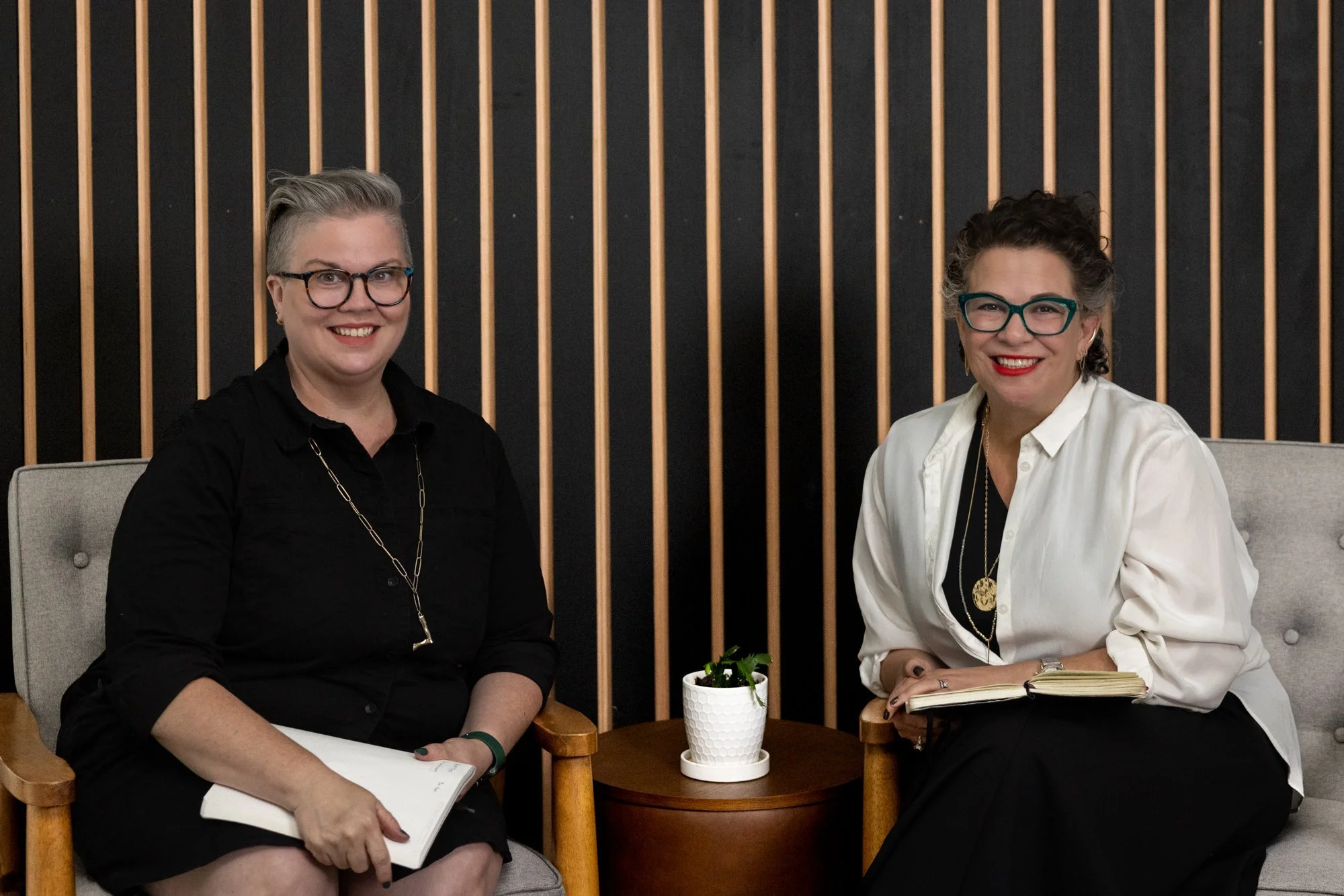 Lora Cover and Sarah Braver, leaders of coLeague, sit in front of a striped background as they both smile brightly at the camera.