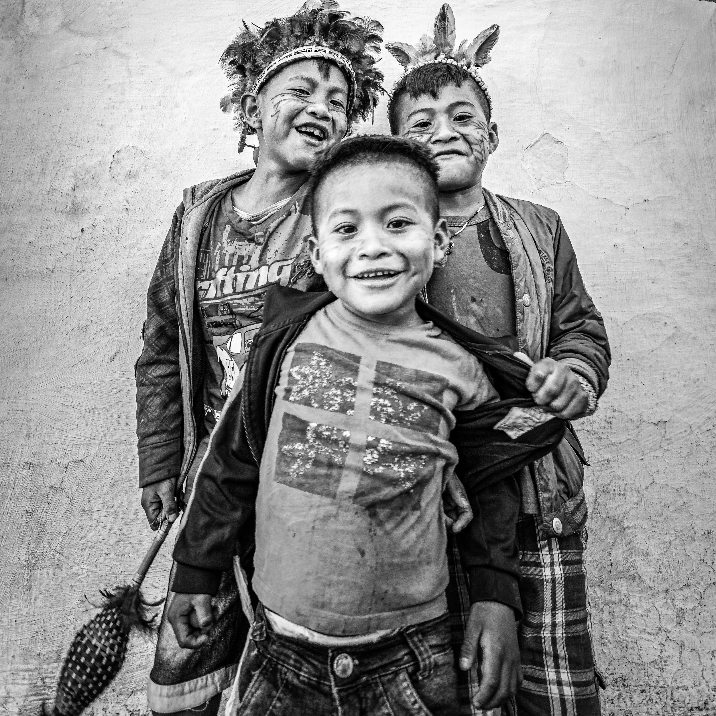 Three smiling children standing in front of a plain wall, with face paint and casual clothes, one holding a small object.