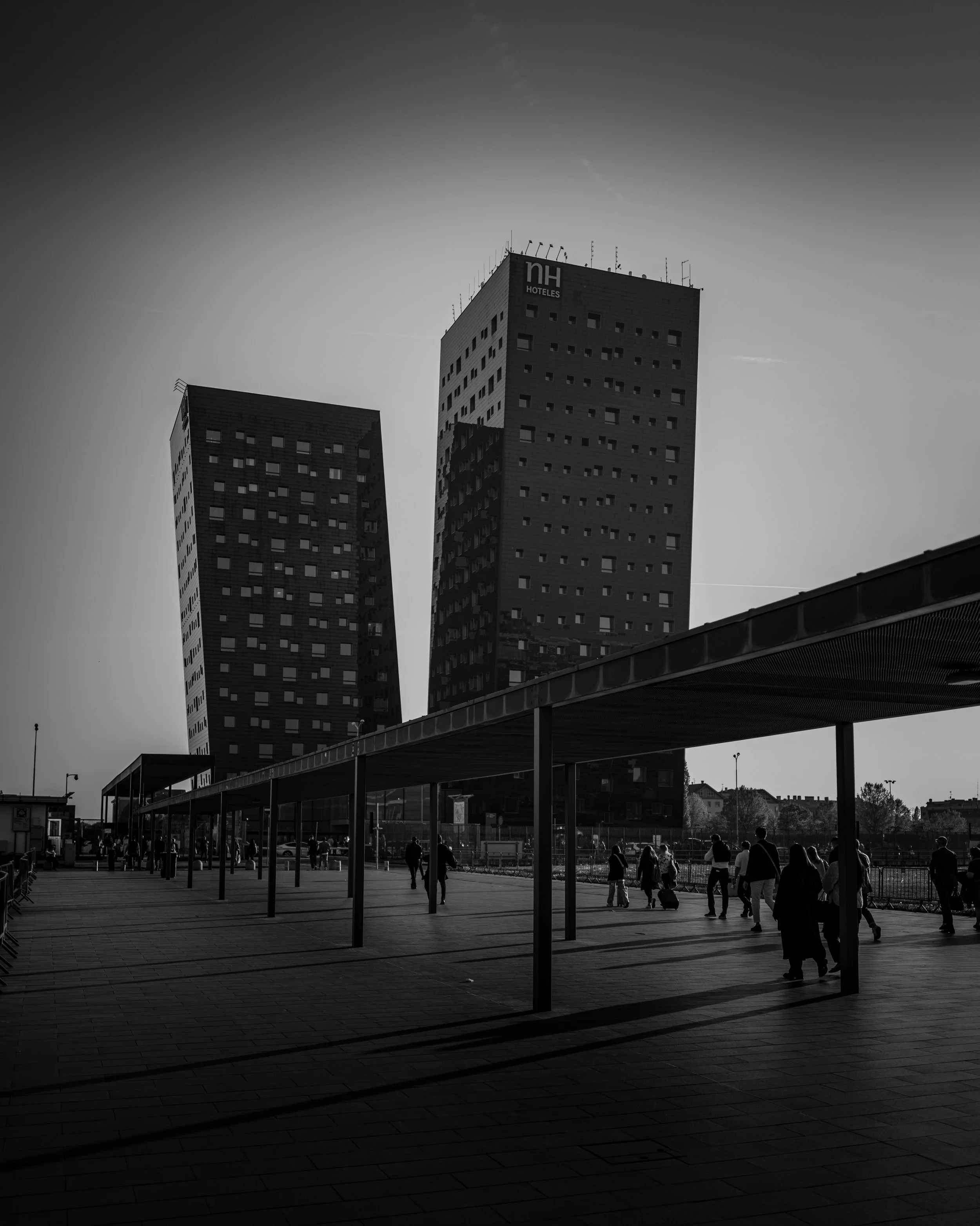 Black and white photo of a cityscape with two modern high-rise buildings and people walking in an outdoor area.